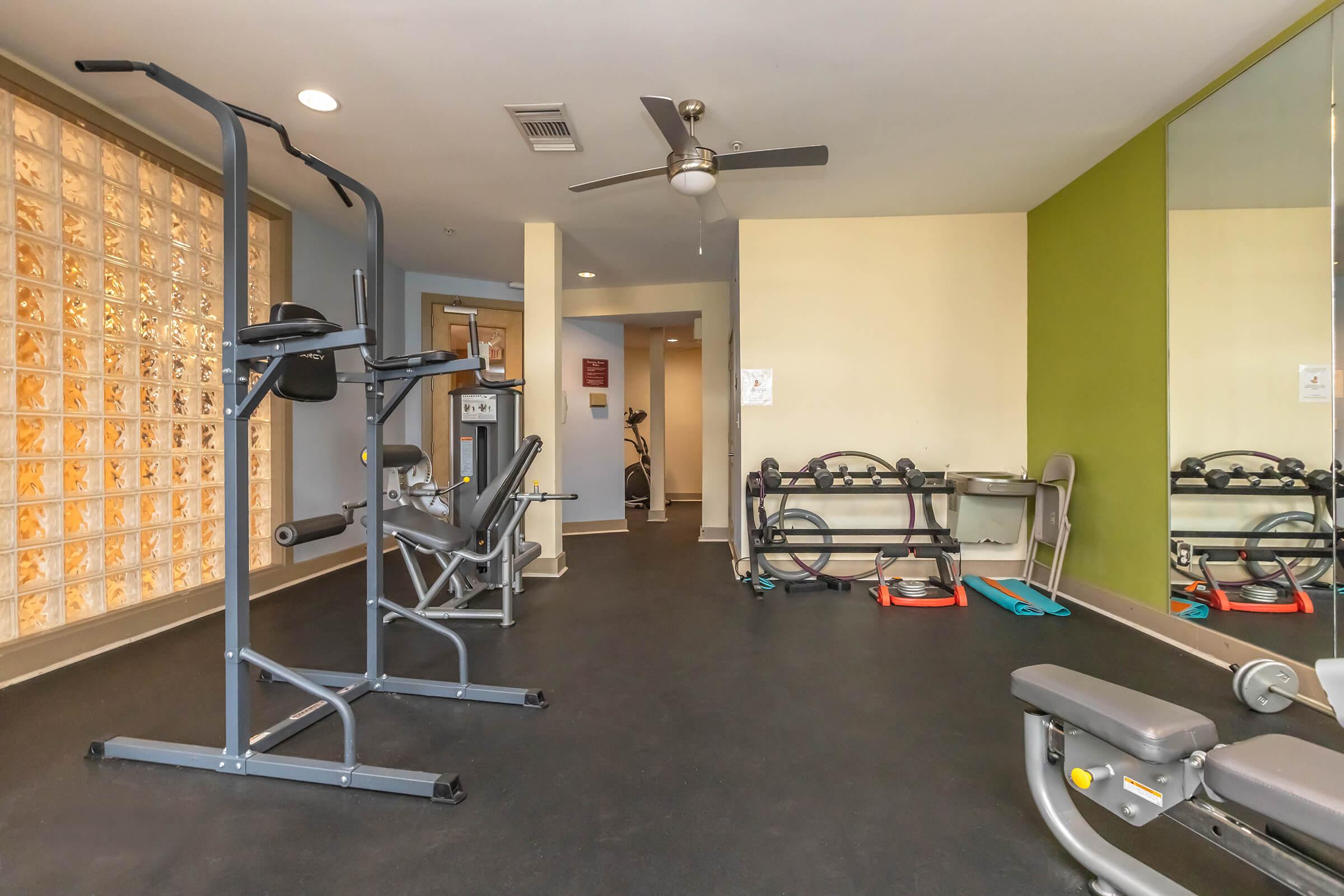 A spacious gym area featuring exercise equipment including a weight machine and a bench press. The walls are painted in calming colors, with a glass block wall providing natural light. A mirror reflects the workout space, and a bicycle is visible in the background.