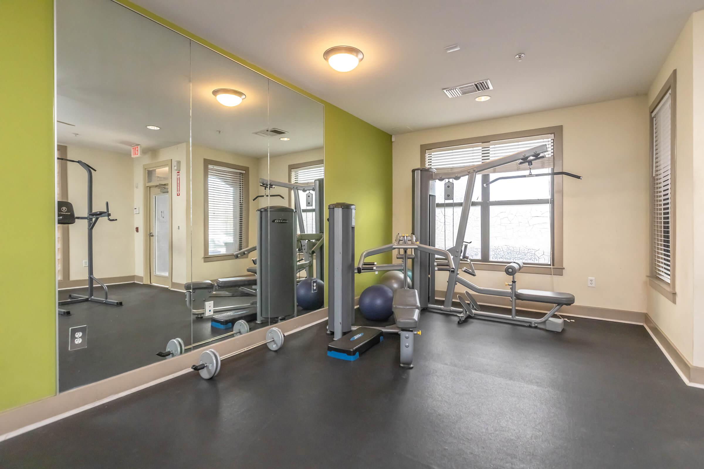 A small gym area featuring exercise machines, free weights, and a stability ball. Mirrors reflect the space, and there are green and neutral-colored walls. Natural light comes in through a window, creating a bright and inviting workout environment.