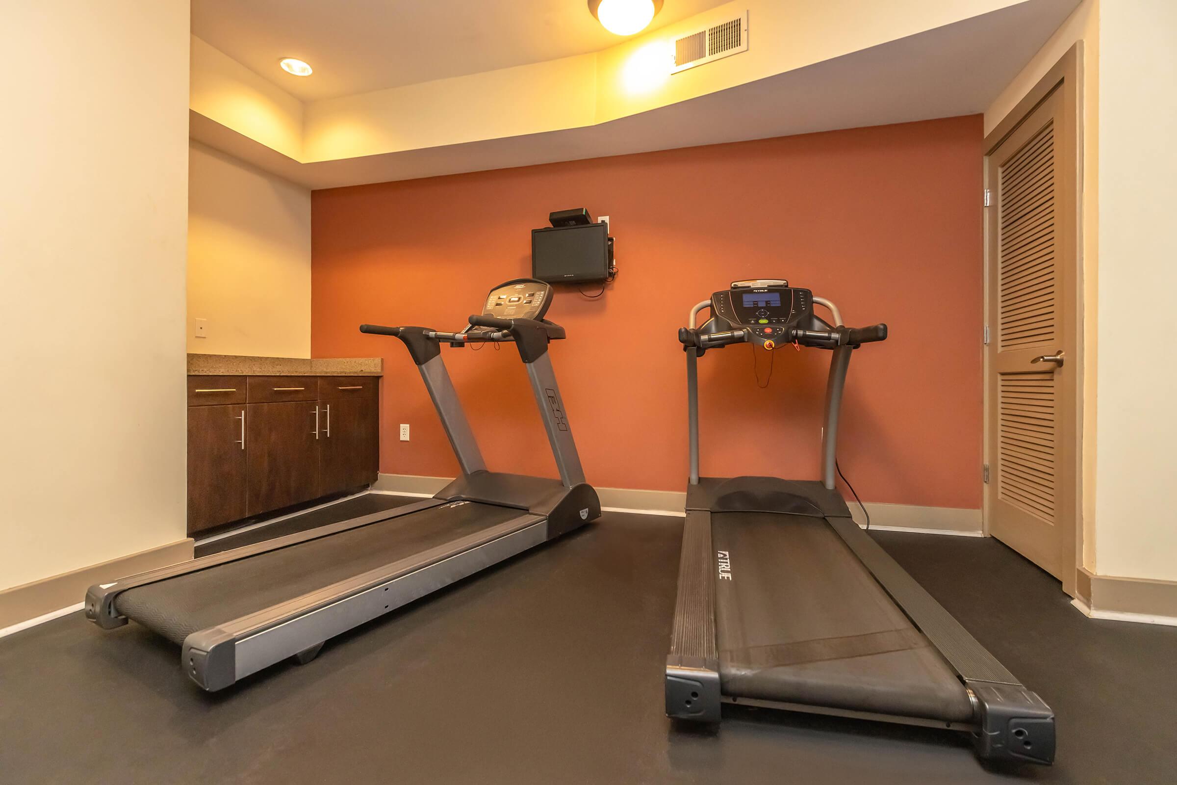 Two treadmills positioned in a fitness space with an orange wall. One treadmill is equipped with a display panel, while the other has a smaller monitor. There is a cabinet with drawers in the background, and a flat-screen TV mounted on the wall. The floor is dark in color, creating a modern workout environment.
