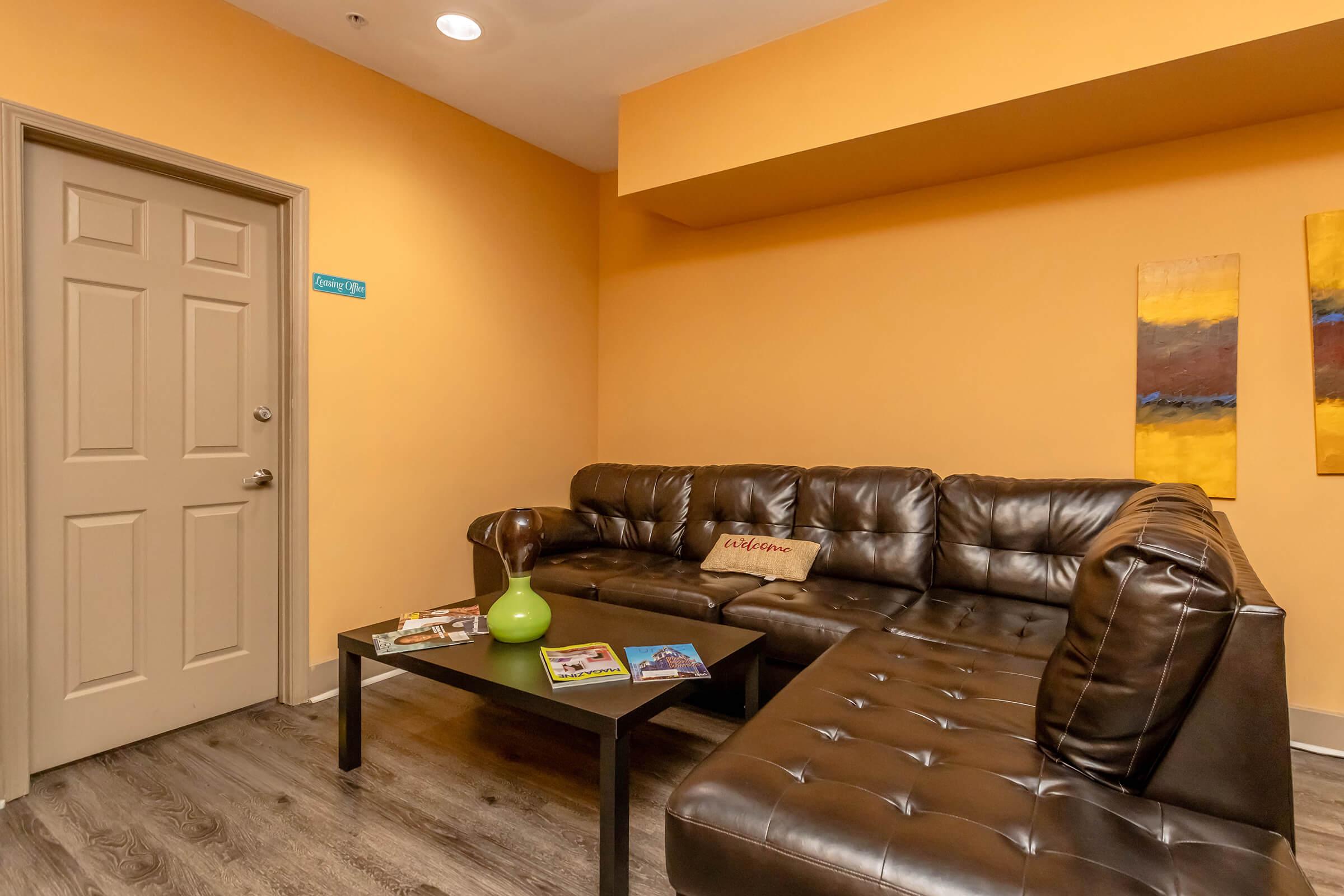 Cozy living space featuring a brown leather sectional couch with decorative pillows. A black coffee table with magazines and a green vase sits in front of the couch. The walls are painted bright orange, and there is a door on the left leading to another room. Two abstract paintings adorn the wall.