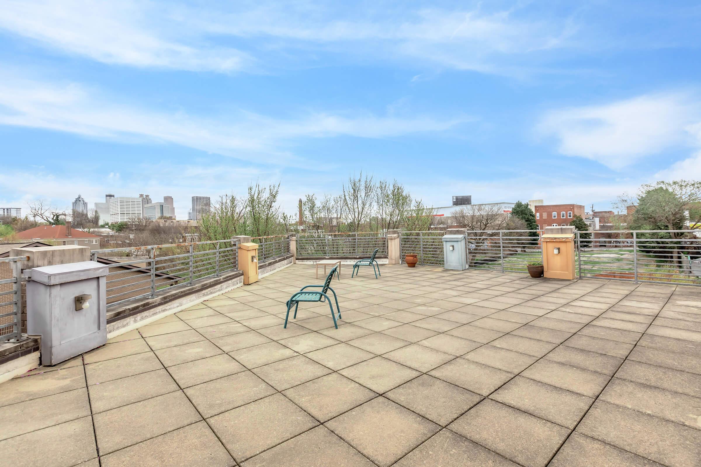 A spacious rooftop terrace with two metal chairs, planters, and trash bins. The area is surrounded by a railing and features a view of distant buildings and trees under a clear blue sky. The floor is paved with large concrete tiles, creating a modern urban outdoor space.