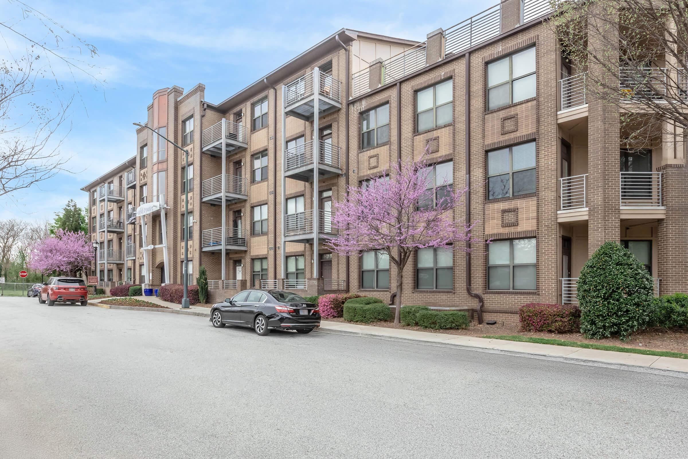 A view of a multi-story brick apartment building with several balconies. In front of the building, there are pink flowering trees and neatly trimmed shrubs. A parked car is visible on the street, along with another vehicle further down. The sky is clear with a light blue hue.