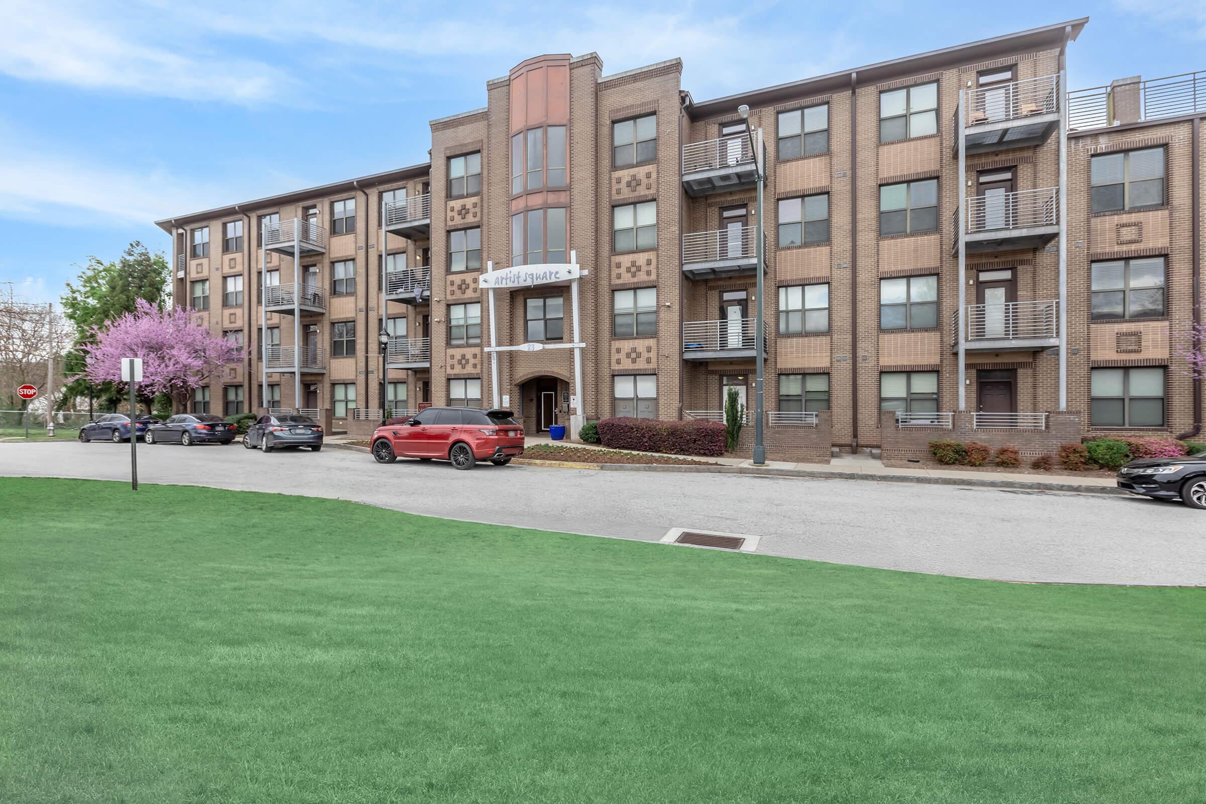 A multi-story brick apartment building with a modern design, featuring a central entrance and several balconies. The surrounding area includes well-maintained grass, trees with pink blossoms, and parked cars along a curved road. The sky is clear and blue, creating a welcoming atmosphere.