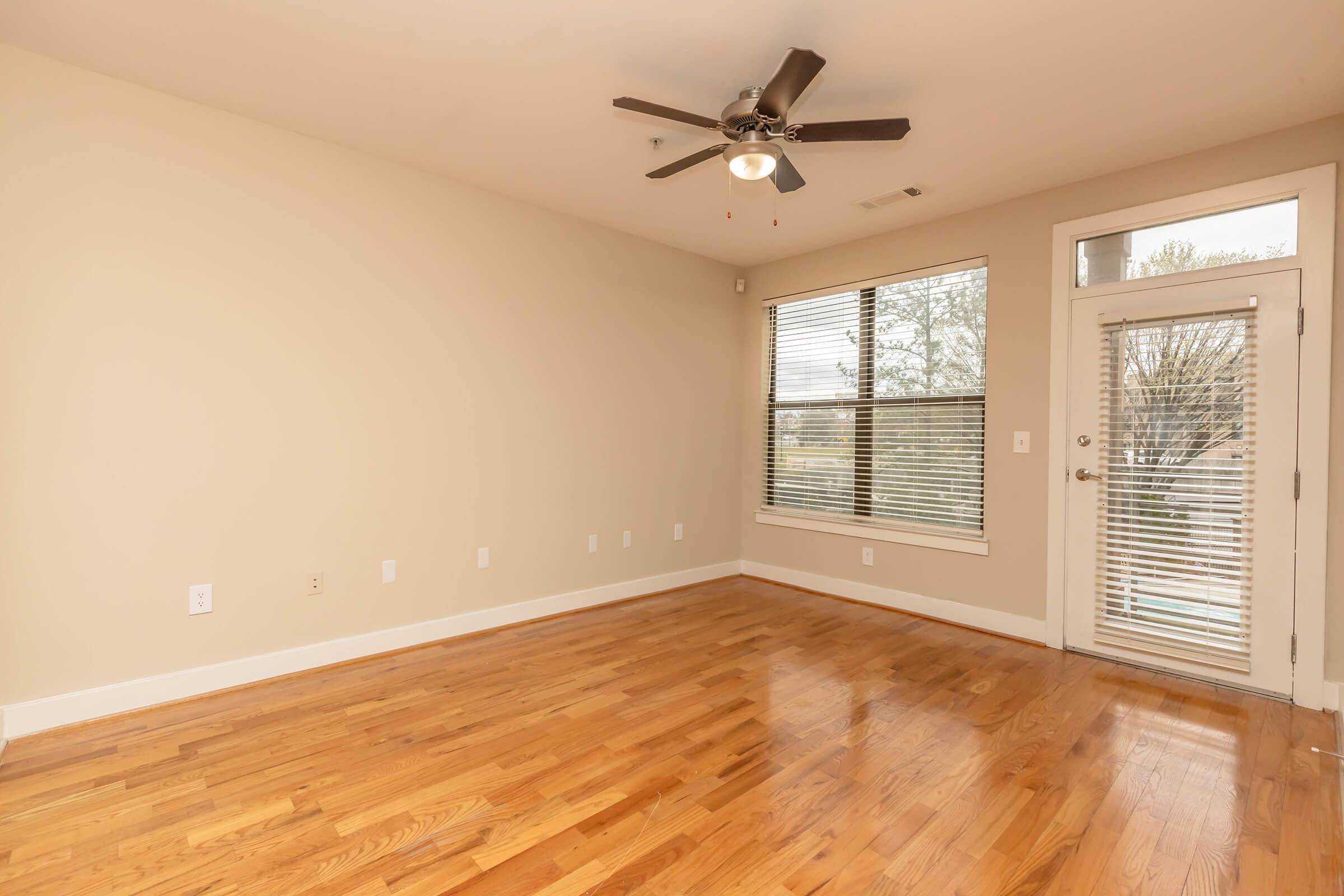 A spacious, empty room with light wood flooring, a ceiling fan, large windows allowing natural light, and a door leading outside. The walls are painted a neutral color, creating a bright and airy atmosphere. Ideal for decorating or furnishing as a living space.