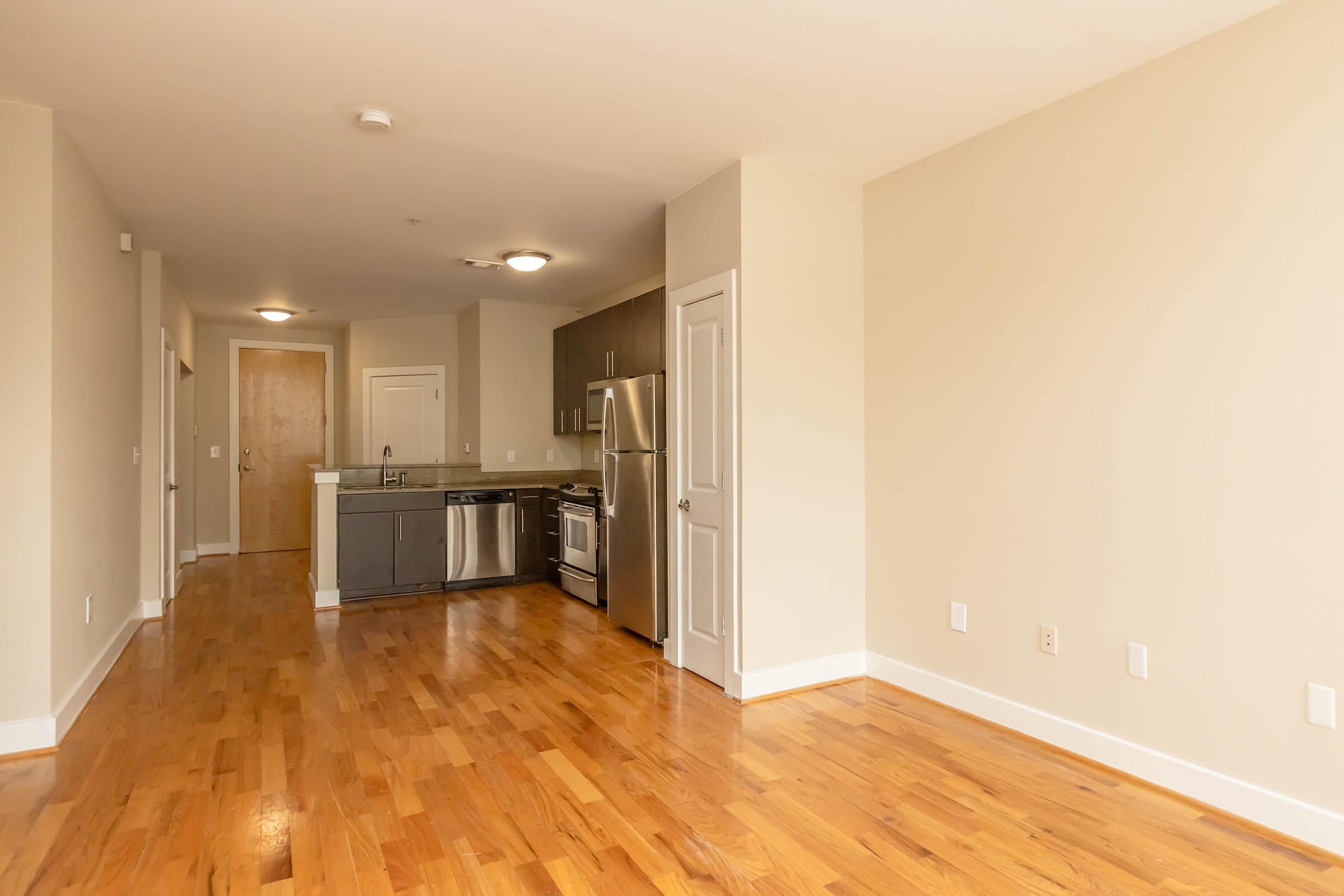 A spacious, well-lit apartment featuring hardwood floors and neutral-colored walls. The room includes a modern kitchen with stainless steel appliances, a refrigerator, and cabinetry. Doorways lead to other areas, and the layout suggests an open-plan design.