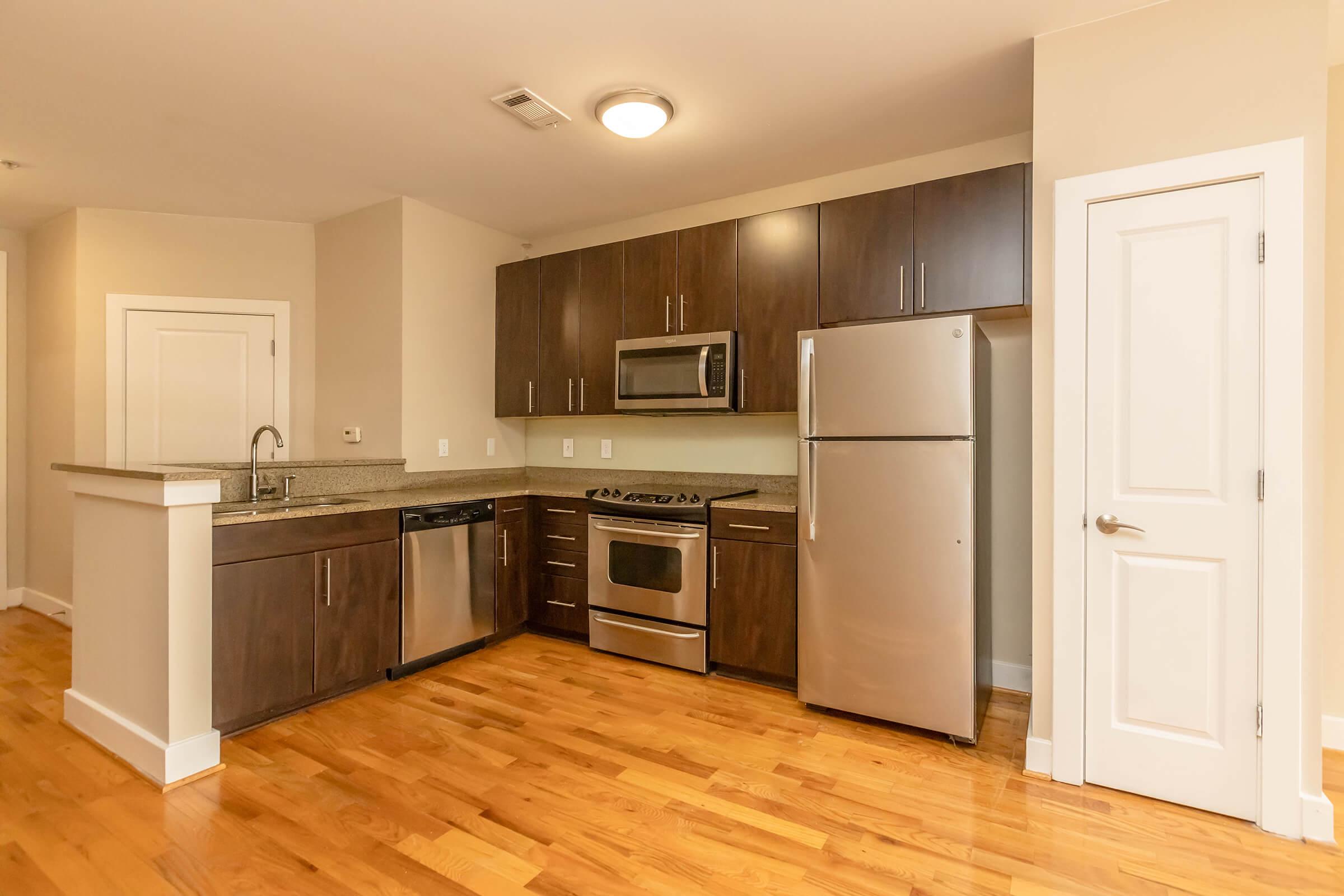 A modern kitchen featuring dark wood cabinetry, stainless steel appliances including a refrigerator, stove, and microwave, a granite countertop, and hardwood floors. A small island separates the kitchen from the living area, with a doorway leading to another room visible on the right.