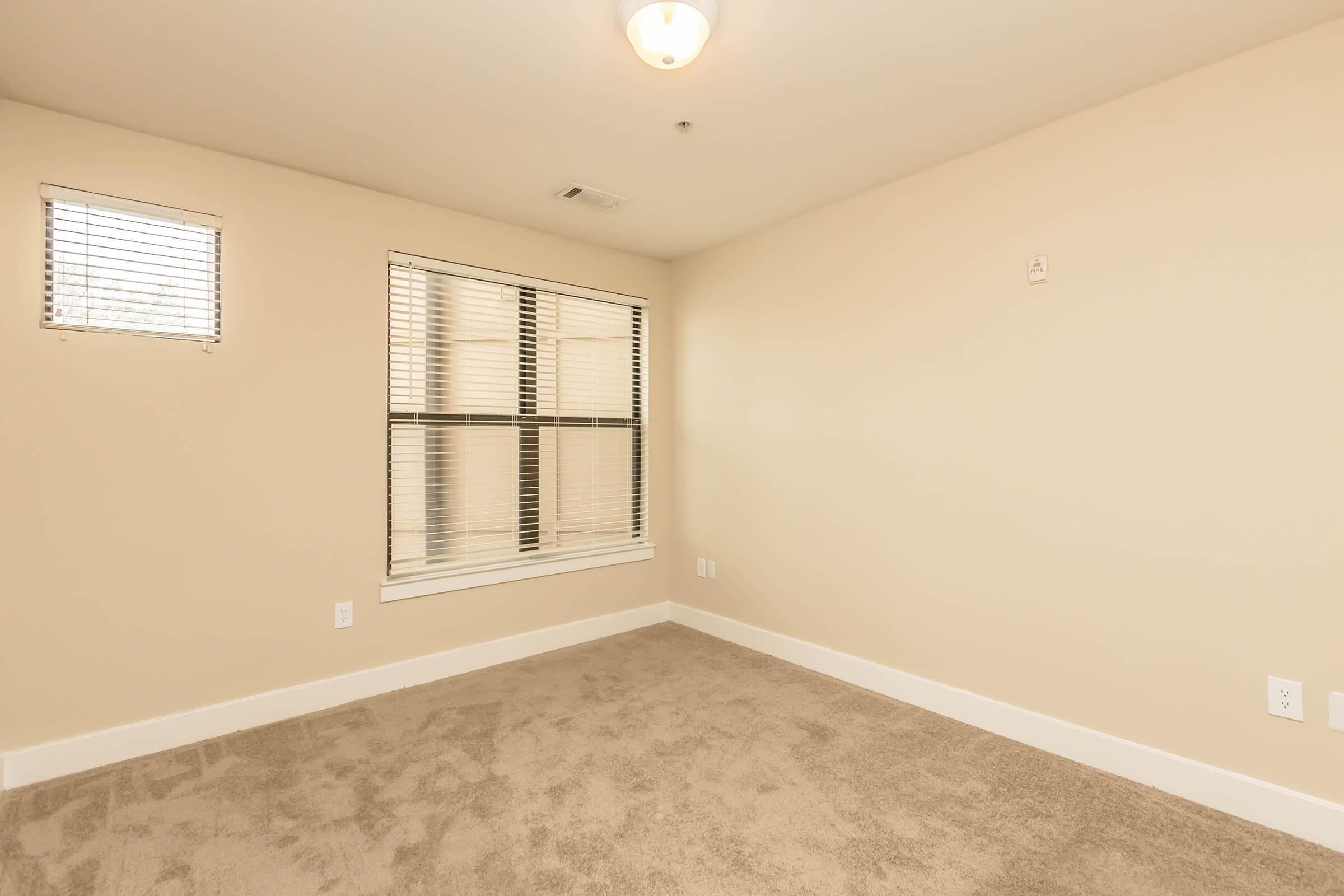 A vacant room with light beige walls, carpeted floor, and a ceiling light fixture. There are two windows with blinds on one wall, letting in natural light, while the opposite wall is plain with no decorations. The space appears clean and ready for furnishings.