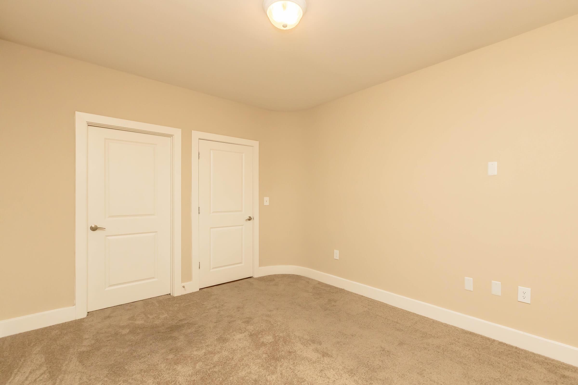 A minimalistic room with beige walls and carpet flooring. Two closed white doors are visible on the left side, and there is a light fixture on the ceiling. The space is empty, creating a blank canvas for potential furniture or decor.