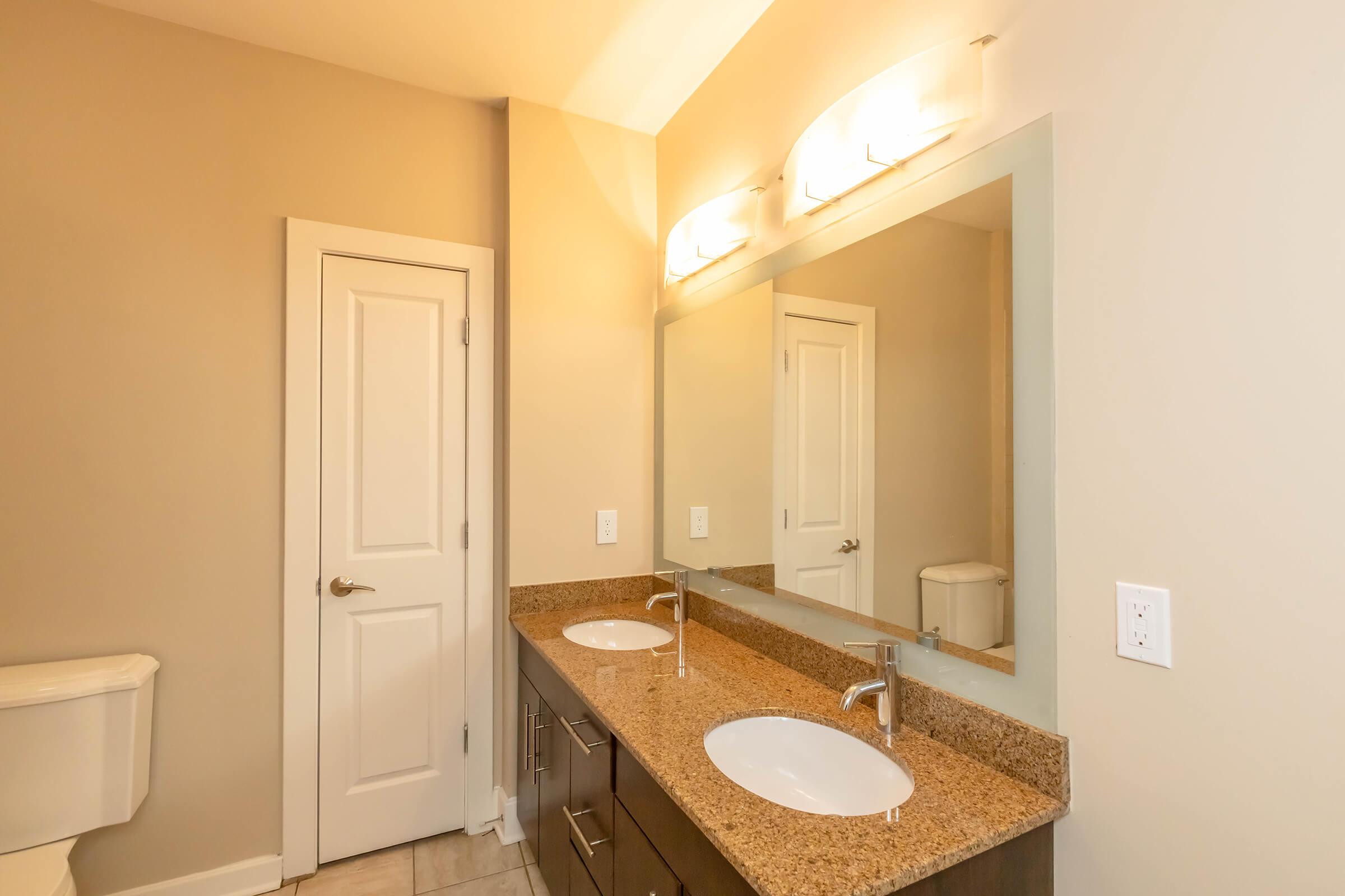 Modern bathroom featuring a double sink vanity with granite countertop, large mirror, and stylish lighting. A white toilet is partially visible to the left, and a closed door is on the right. The walls are painted in a soft beige color, creating a warm and inviting atmosphere.