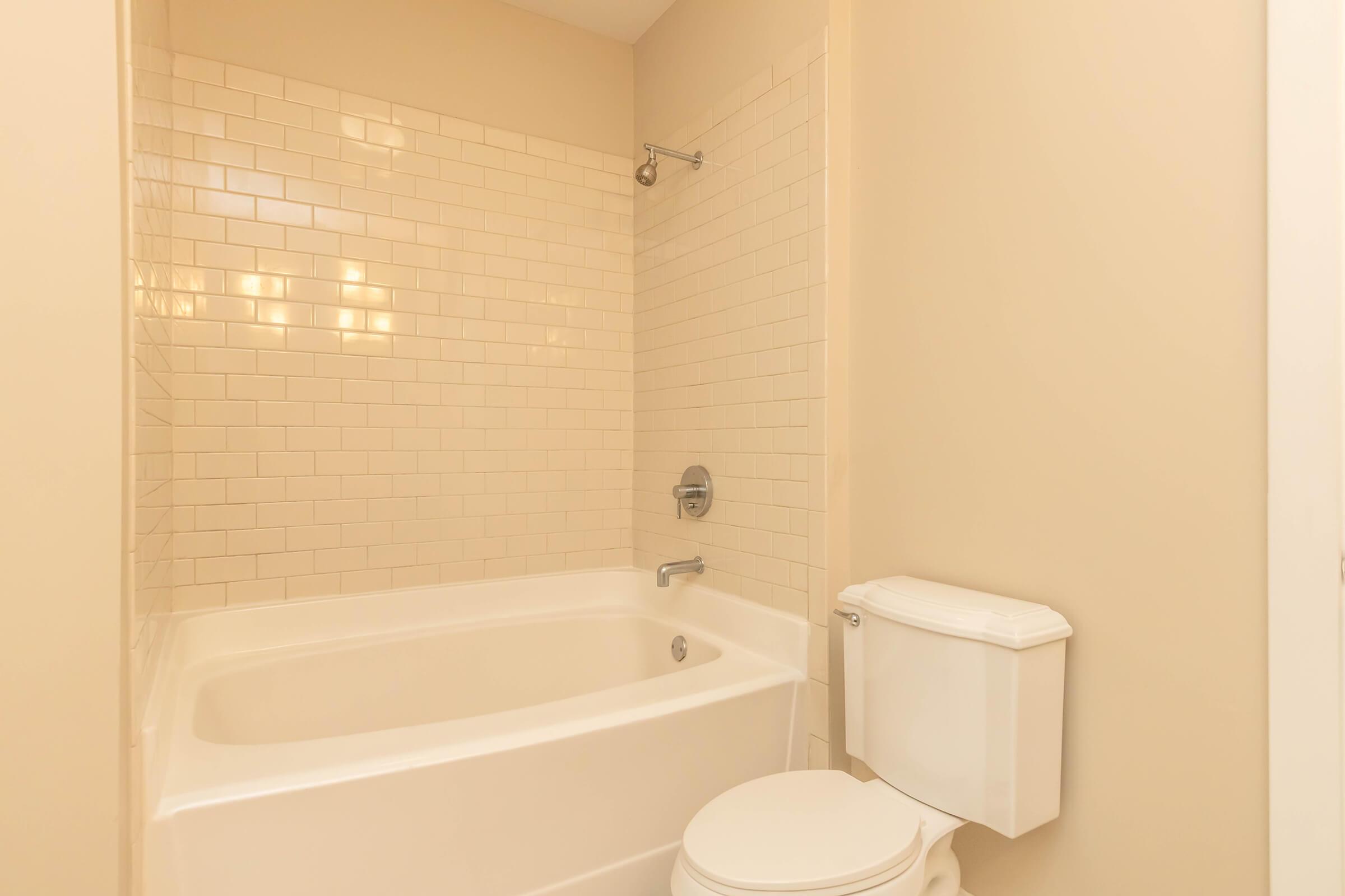 A clean and neutral-colored bathroom featuring a bathtub with a showerhead, a toilet, and beige tiled walls. The space has a minimalistic design with good lighting.
