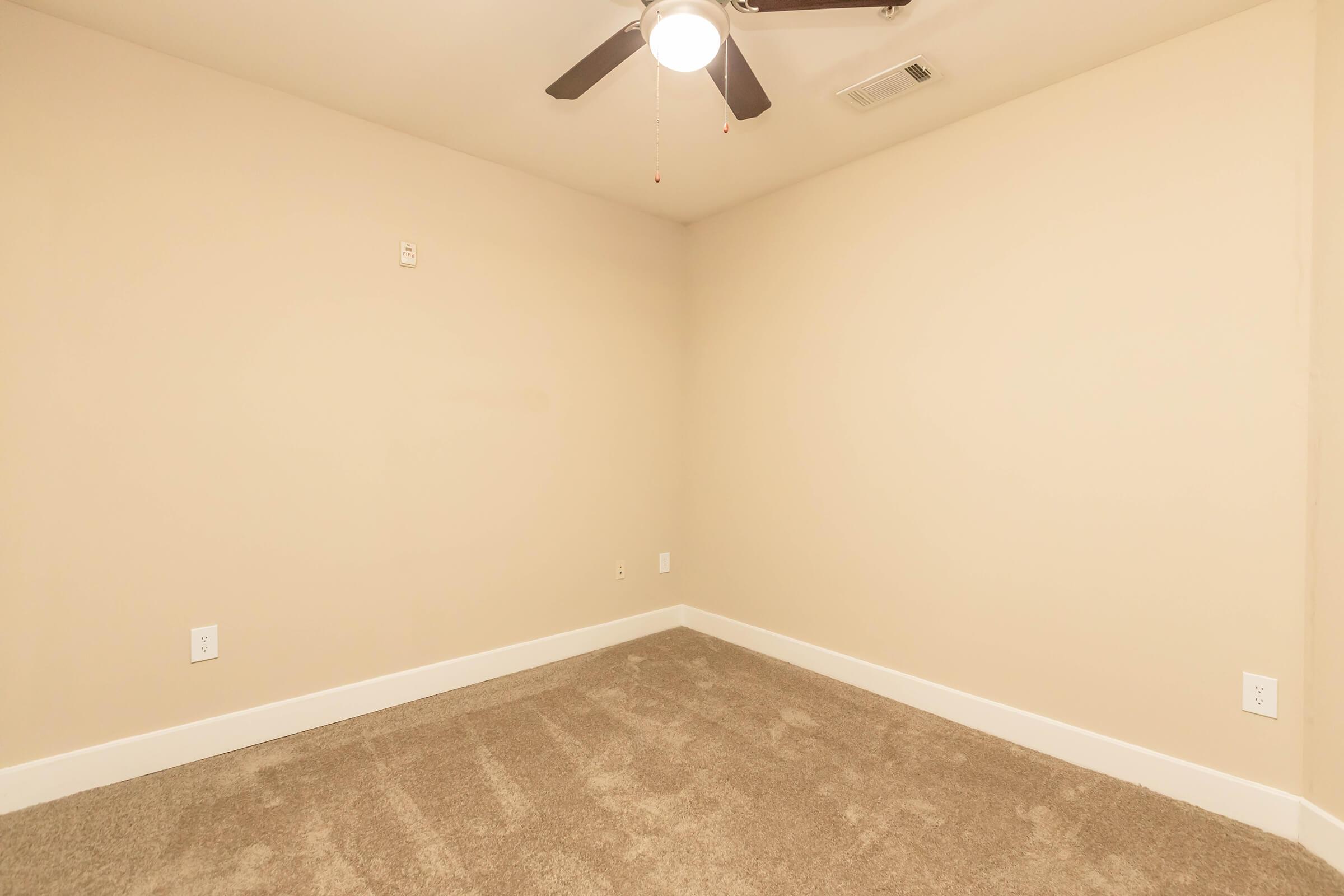 Empty room with beige walls and a ceiling fan. The floor is covered with carpet, and there are light switch panels on the walls. The space appears fresh and ready for decoration or furniture placement.
