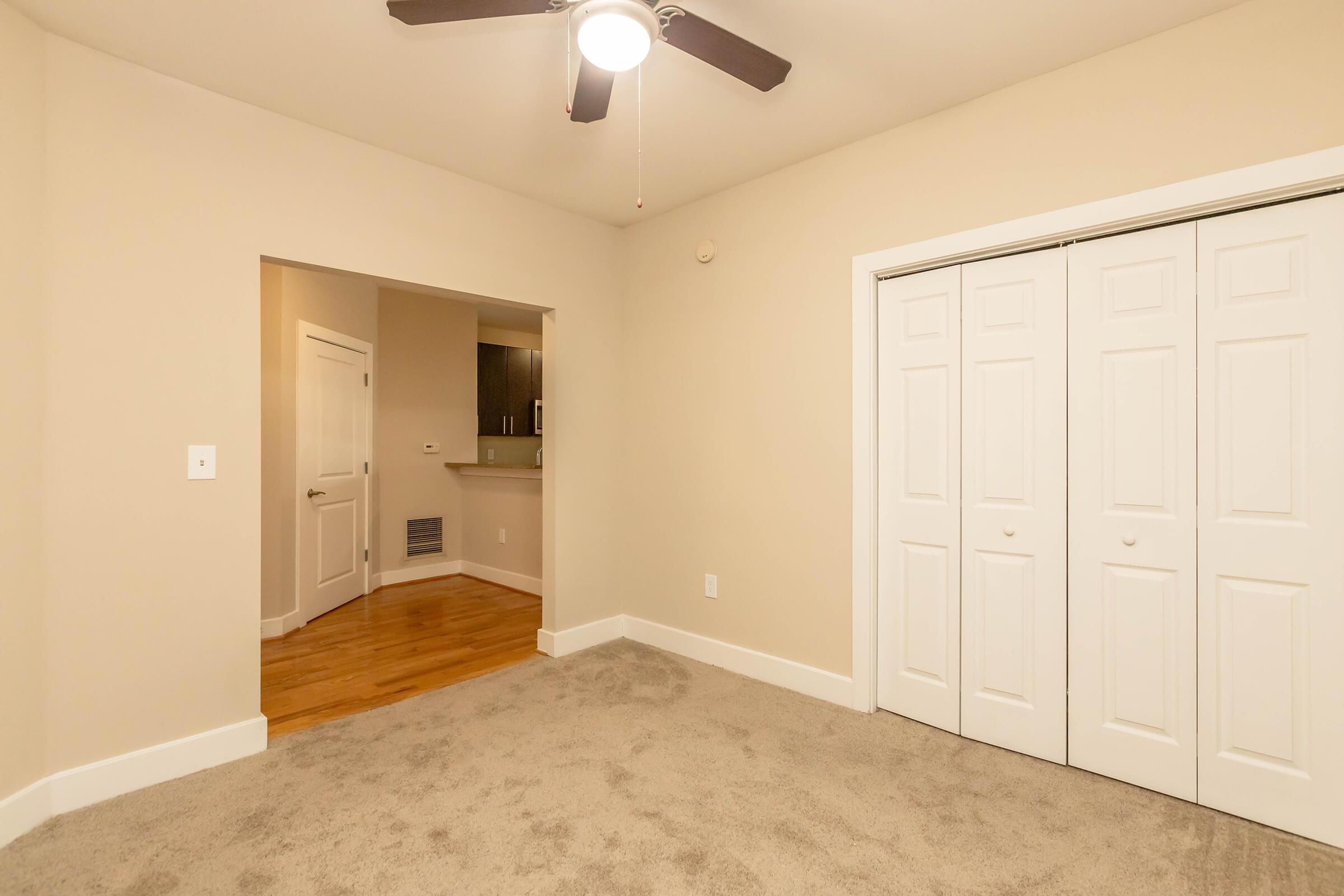 A spacious, well-lit room with beige walls and carpeted flooring. It features a ceiling fan and a double closet with sliding white doors. An open doorway leads to a kitchen area with modern cabinetry, visible in the background. The overall ambiance is inviting and suitable for relaxing or living.