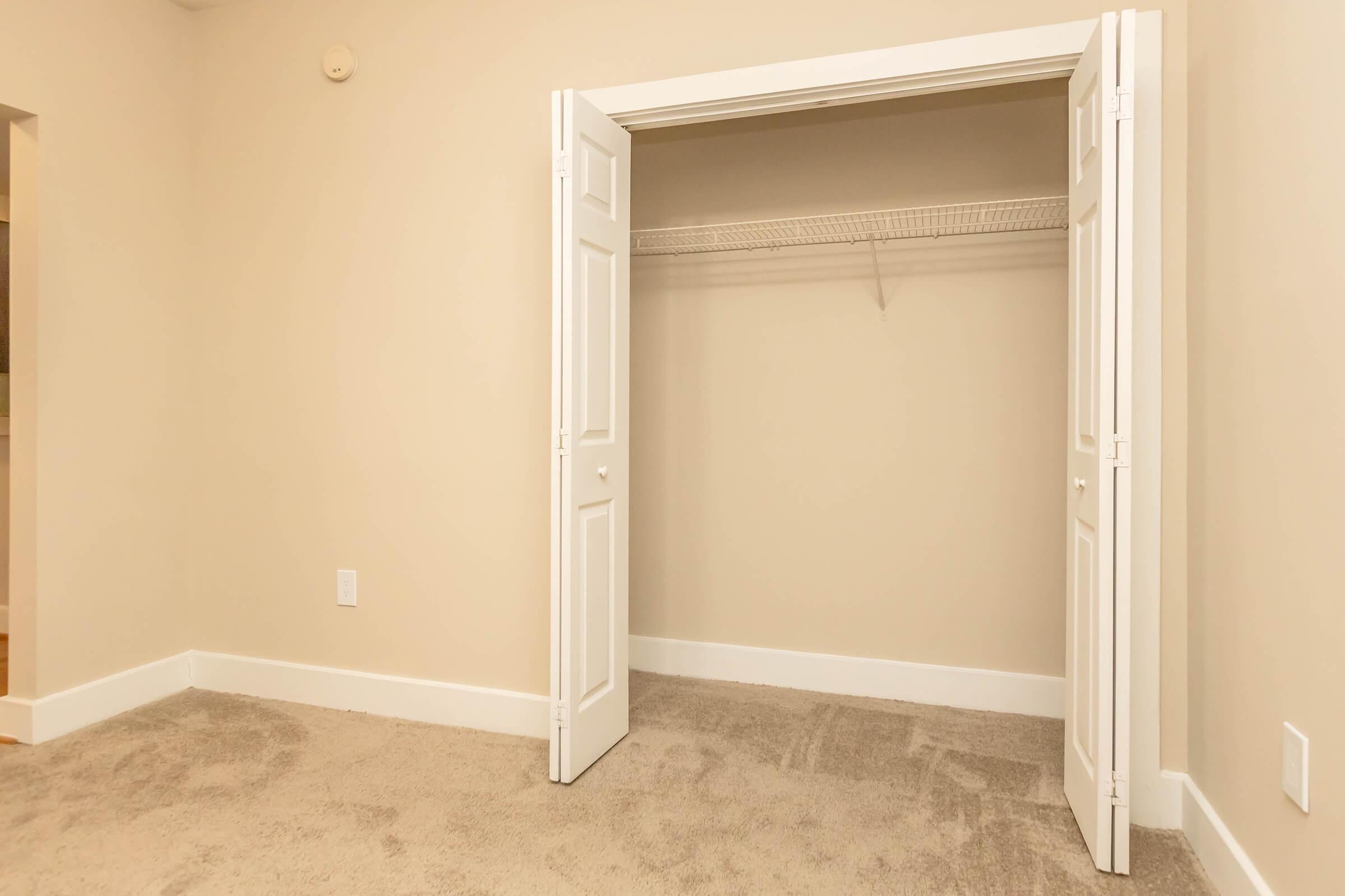 Empty closet with double white doors, showcasing a beige wall and a single shelf inside. The floor has light beige carpet, and there's a small white electrical outlet visible on the left wall. The overall space appears bright and clean, suitable for storage.