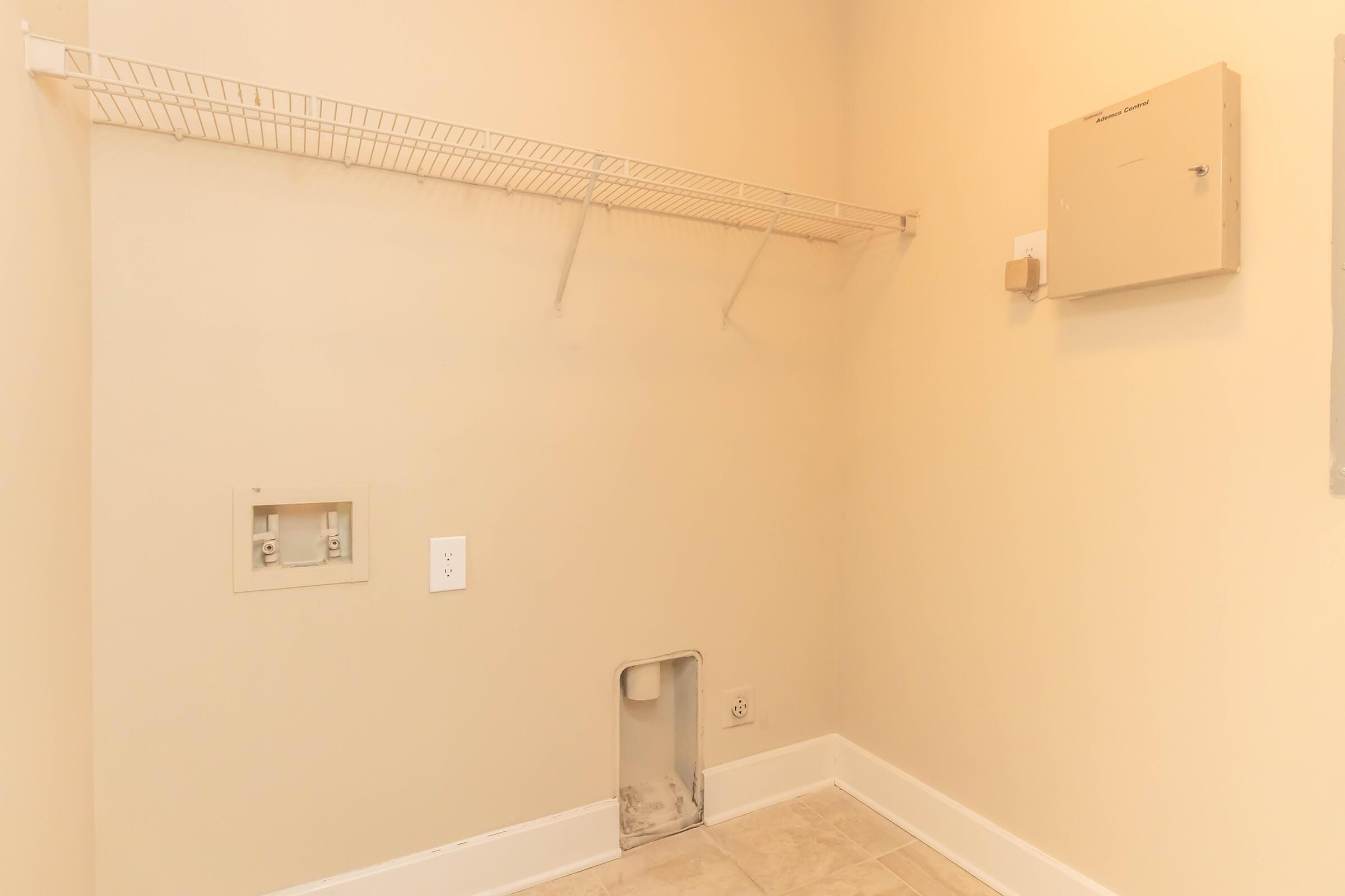 An empty laundry room with cream-colored walls. There is a wire shelf mounted on the wall, and an electrical panel is visible on the right. The floor is tiled, and there is a small opening on the left side, possibly for plumbing or utilities. The space is well-lit and devoid of furniture or appliances.