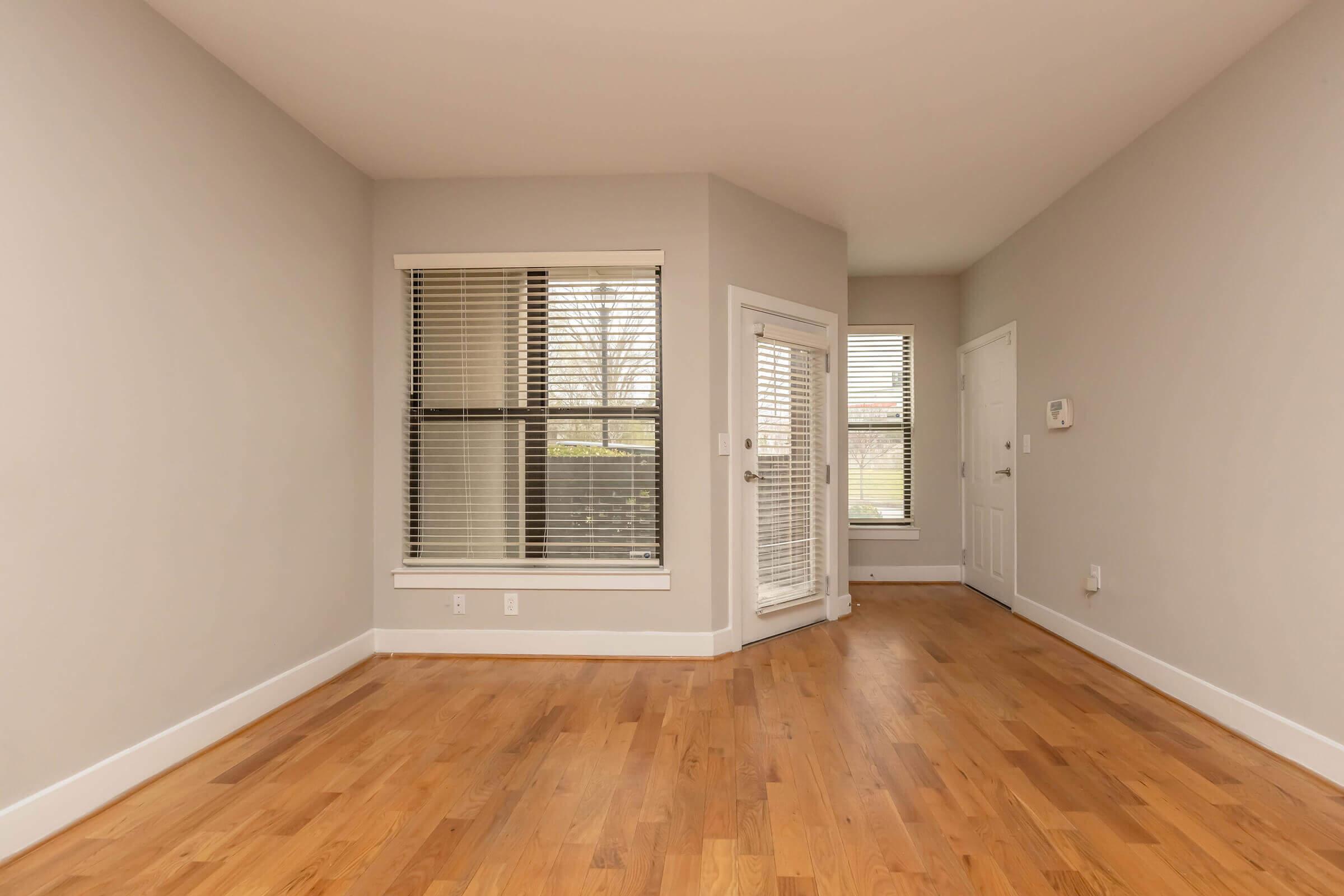 A spacious room with light gray walls and hardwood flooring. It features large windows with blinds, allowing natural light to enter. There is a sliding glass door that opens to an outdoor area, and a small entrance to another room on the right. The overall atmosphere is bright and airy.