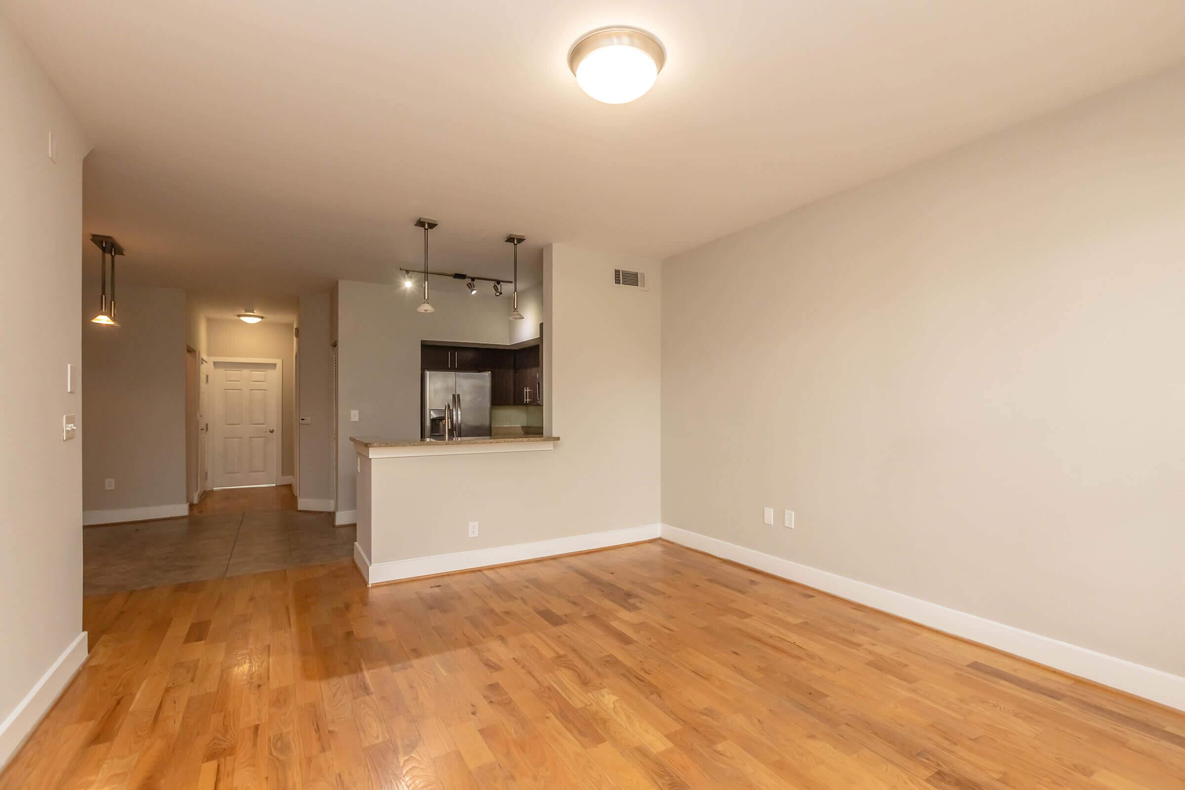 A spacious, neutral-colored living area with hardwood floors. It features a well-lit kitchen area with bar seating, modern light fixtures, and an open layout. The entrance is visible in the background, showcasing a clean, minimalist design.