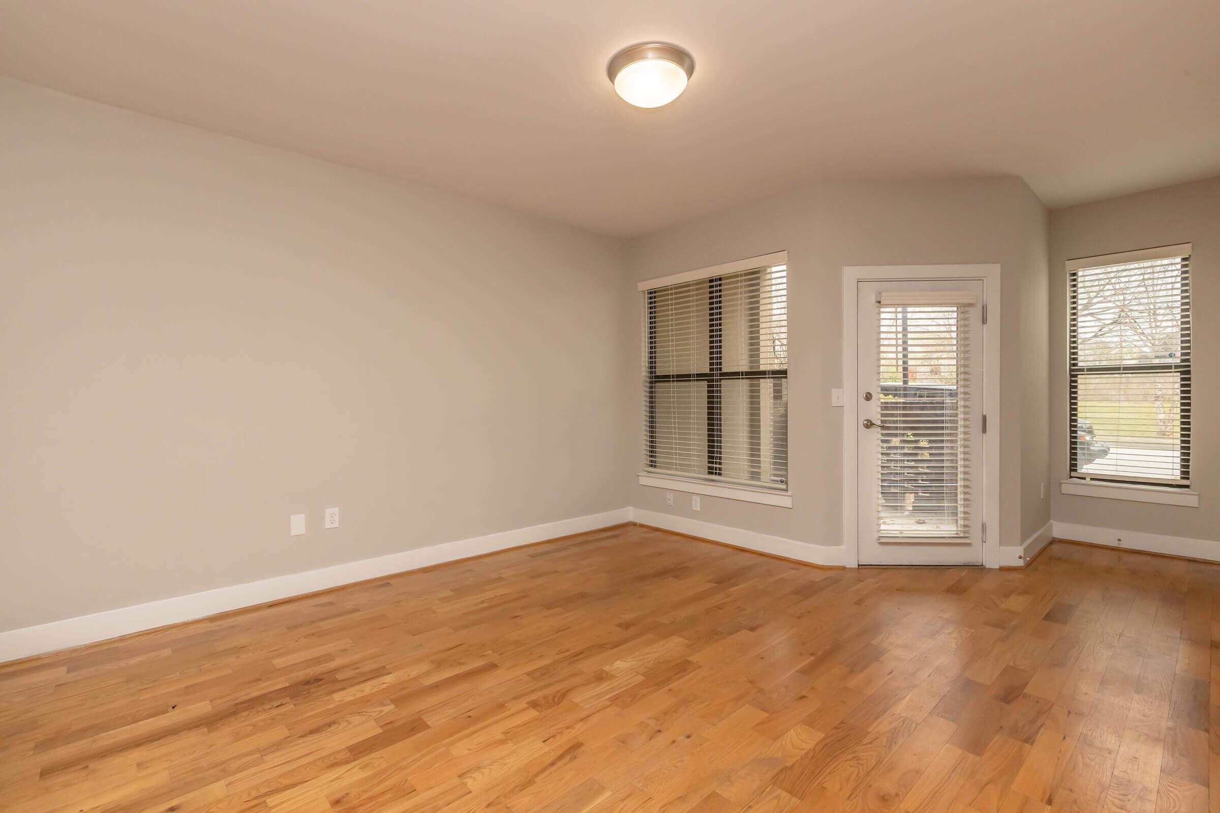A spacious, empty room featuring light-colored walls and hardwood flooring. There are multiple windows allowing natural light, and a door leading outside. The room has a simple ceiling light fixture and no furniture, creating an open and airy atmosphere.