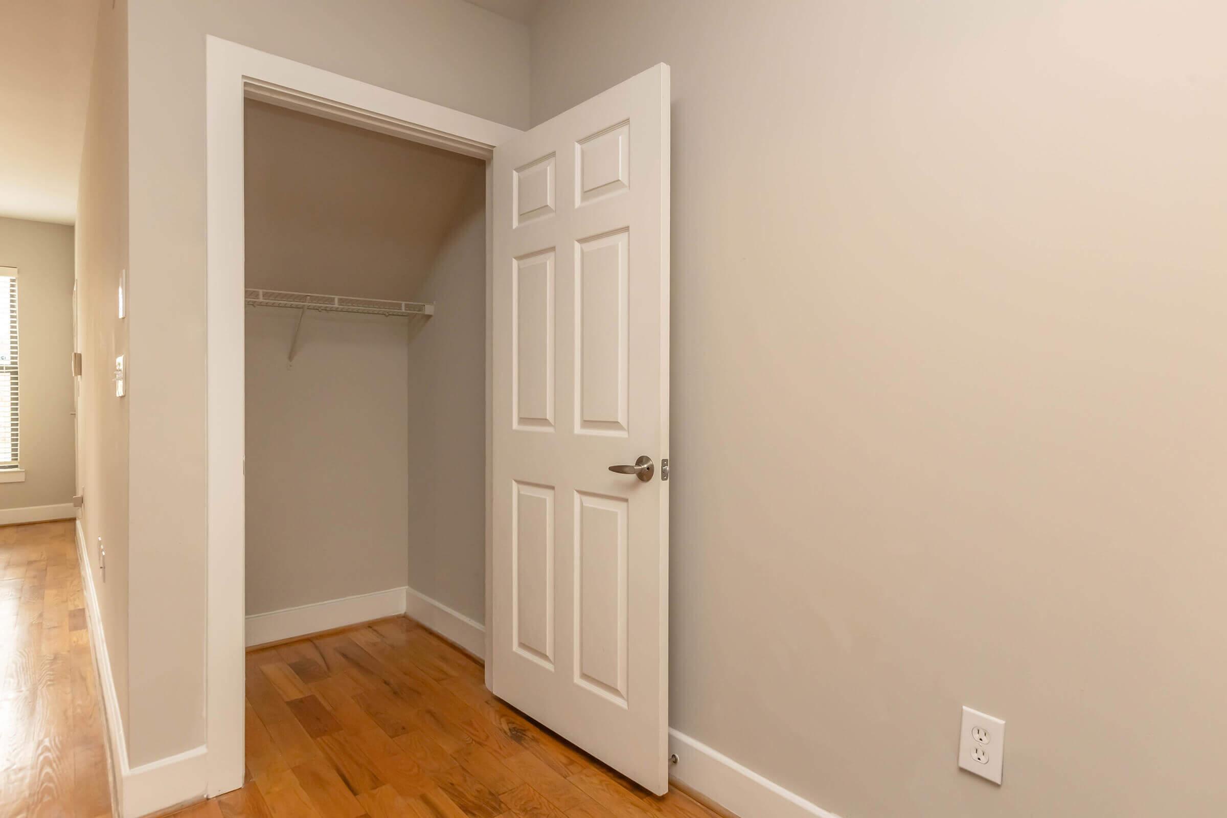 A small, empty room with light gray walls and a wooden floor. On the right, there's a white door slightly ajar, revealing a closet with a shelving unit inside. The space appears bright and minimalistic, showcasing clean lines and a simple design.