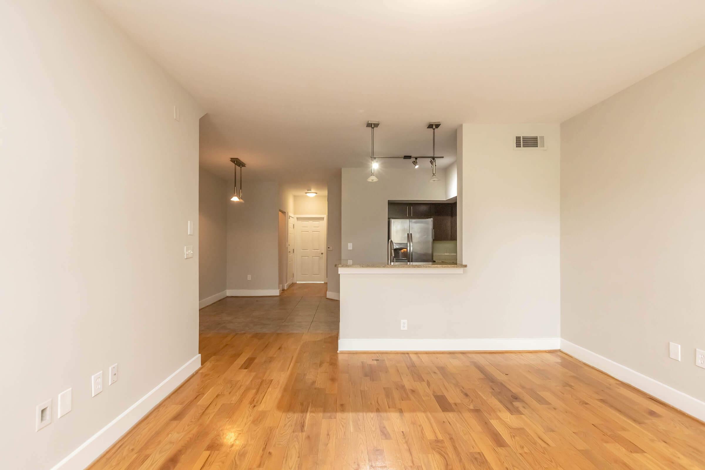 A spacious and modern apartment interior featuring hardwood flooring, light-colored walls, and a kitchen area visible in the background. The room is illuminated by natural light, with recessed lighting and pendant fixtures. The open layout creates a welcoming atmosphere.