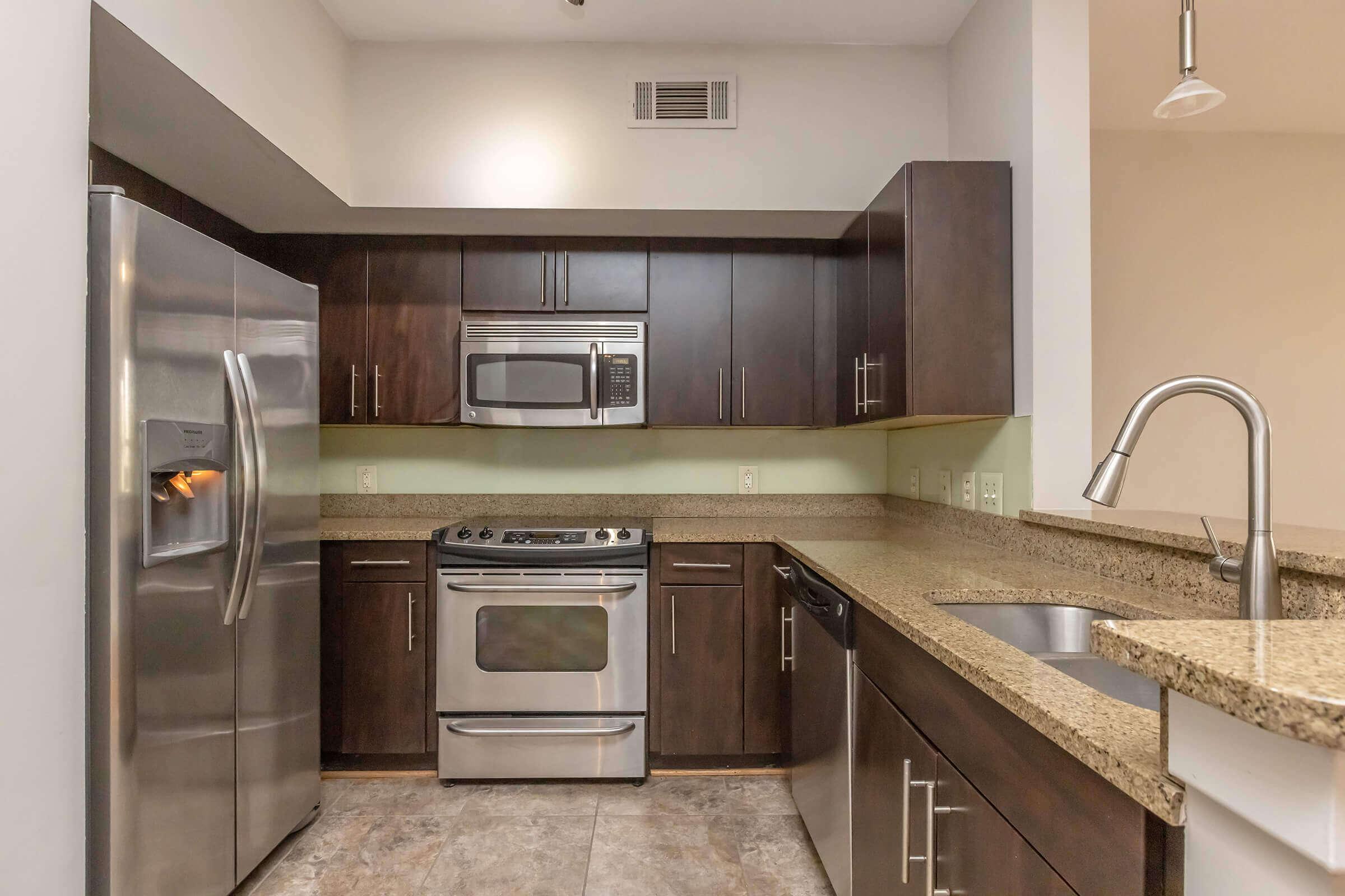 Modern kitchen featuring dark wood cabinets, stainless steel appliances including a refrigerator, microwave, and oven. There is a granite countertop, a double sink, and a light-colored backsplash in a well-lit space. The flooring is a neutral, speckled design.