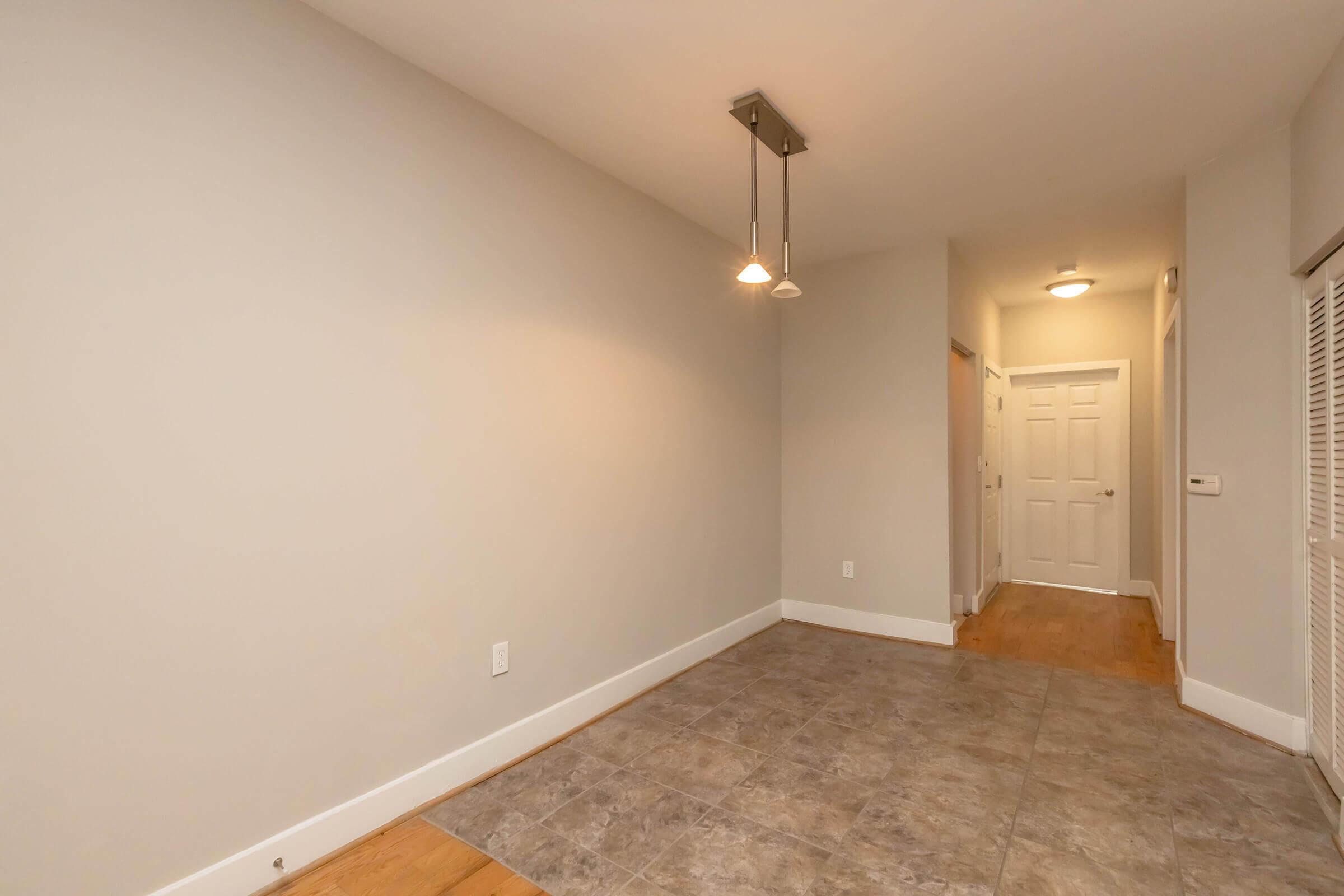 A spacious hallway with light-colored walls and a tiled floor. There are two doors visible: one leading to a room and another closed door further down the hall. A modern light fixture with two bulbs hangs from the ceiling, illuminating the area. The hallway is uncluttered and has a warm atmosphere.