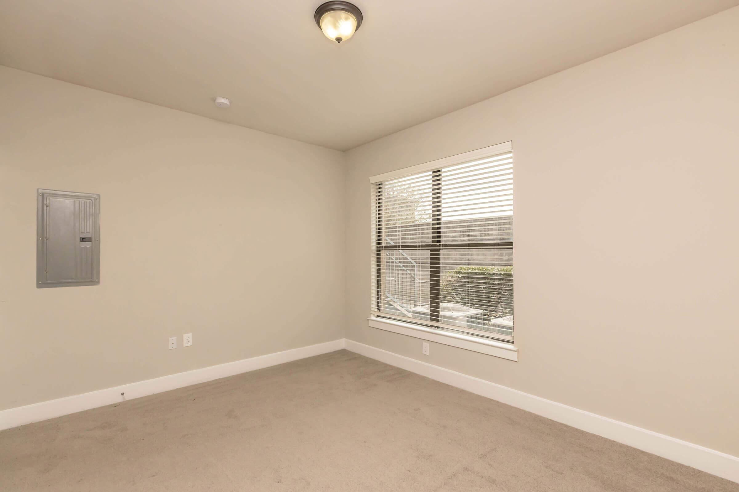 Empty room with light beige walls, a window with blinds overlooking a view outside, and a ceiling light fixture. The floor is covered in light-colored carpet, and there's an electrical panel on one wall. The room has a minimalistic and clean design.