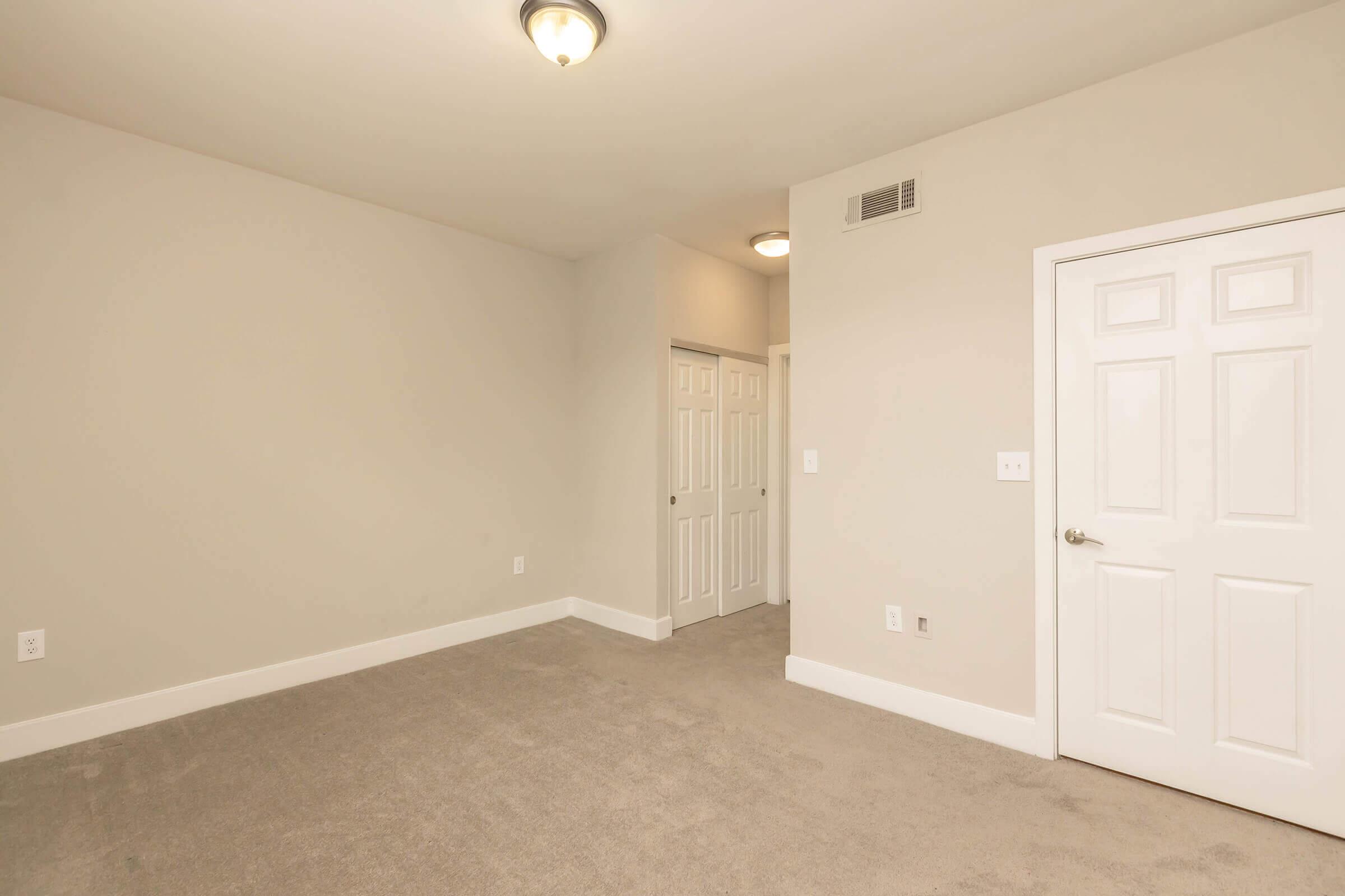Empty room with light beige walls and a carpeted floor. A single overhead light fixture is visible. Two doors are on the right side: one is a standard door and the other appears to be a closet door. The space is bright and uncluttered, giving a sense of openness.