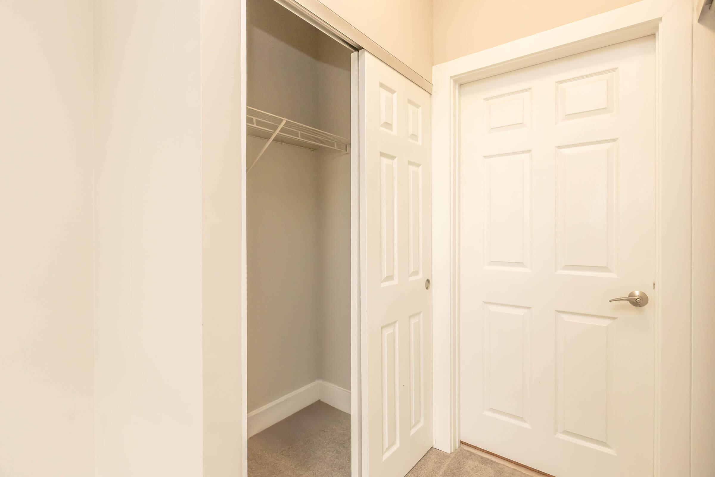 A bright, clean hallway with a white closet door slightly ajar, revealing a simple, empty closet with a hanging rod. Adjacent to it is a closed white door, both doors framed by light-colored walls and a soft carpeted floor, creating a tranquil and unadorned atmosphere.