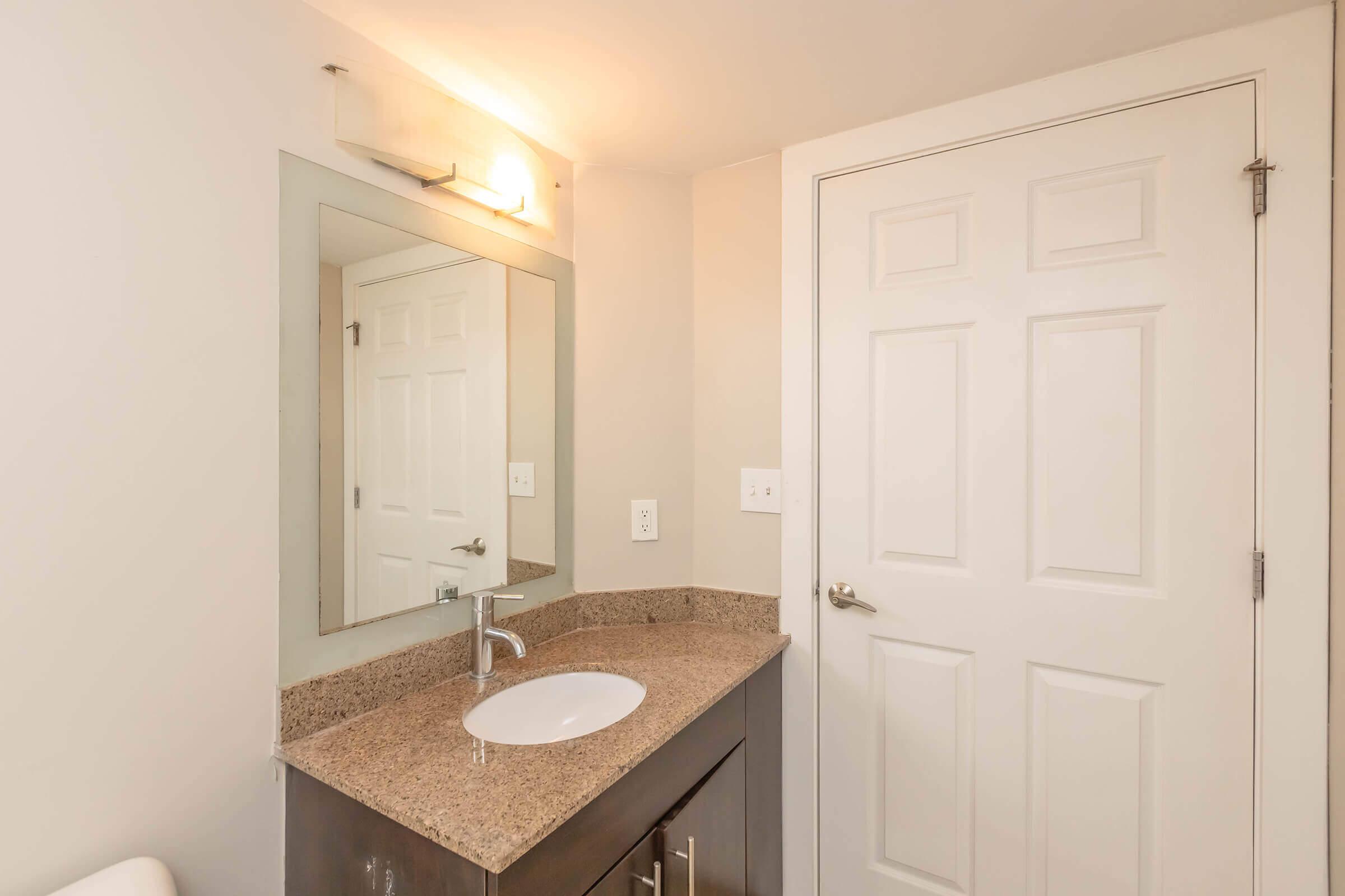 A modern bathroom featuring a granite countertop with a sink, a large mirror above the sink, a light fixture, and a closed white door. The walls are painted in a light color, creating a bright and clean appearance. The overall design is simple and contemporary.