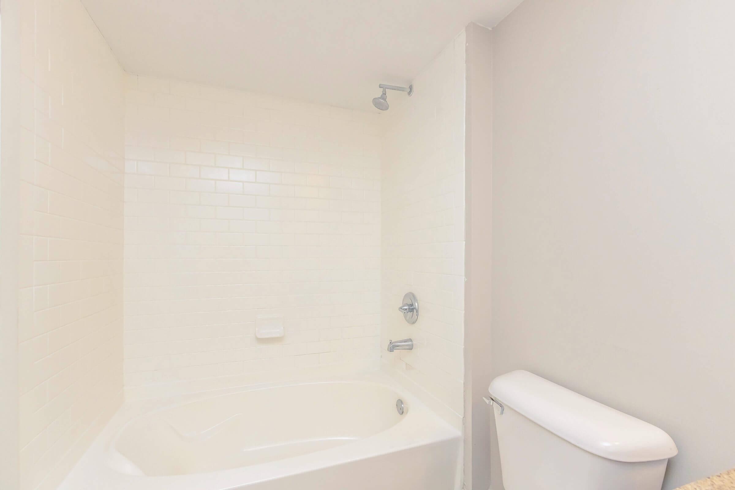 A clean, minimalist bathroom featuring a white bathtub with a tile surround, a chrome showerhead, and a toilet. The walls are light-colored, enhancing the bright and airy feel of the space.