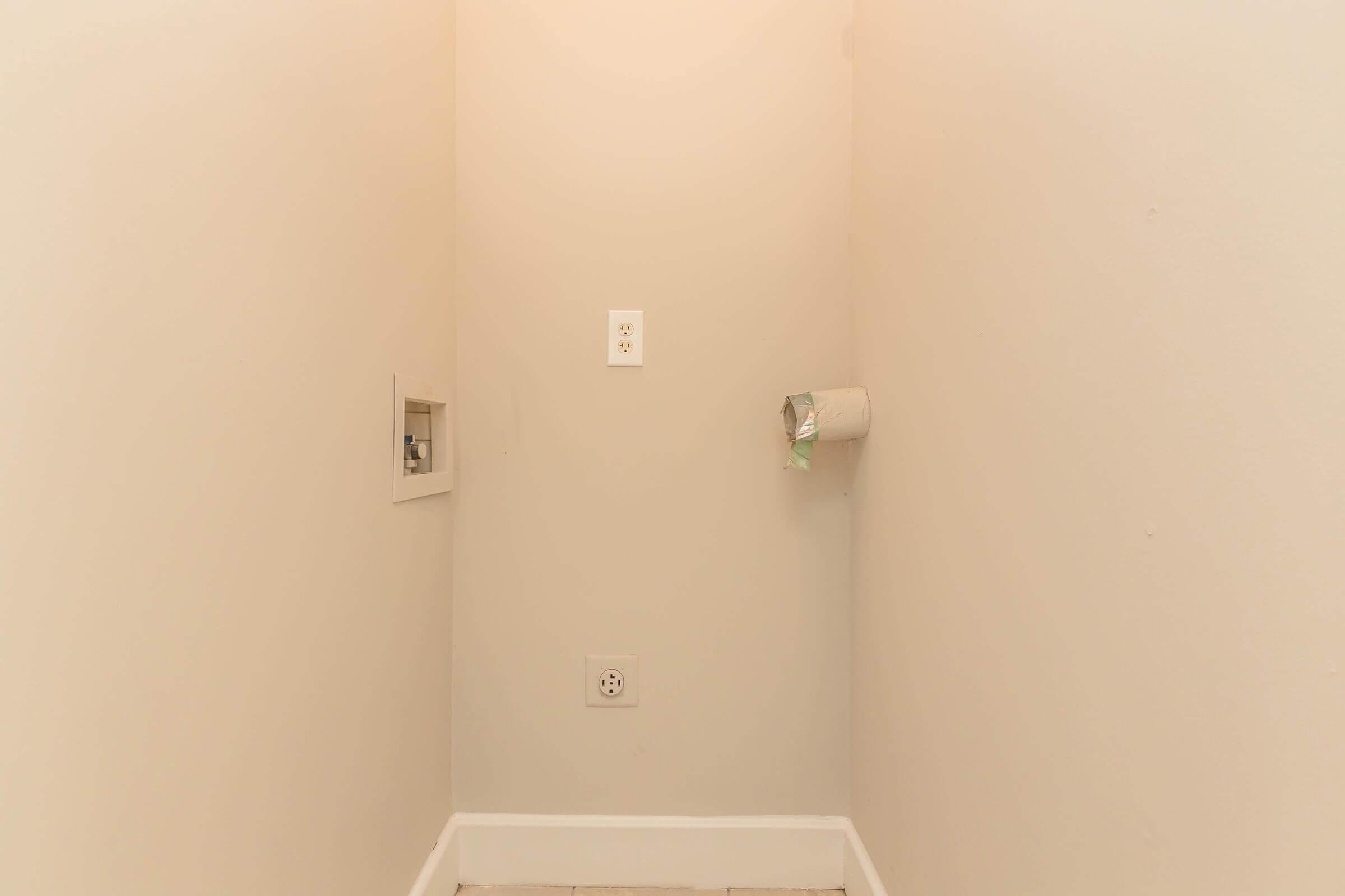A small, empty corner of a room with light beige walls. There is a light switch and an electrical outlet on one wall, and a small wall-mounted fixture for a toilet paper roll visible. The floor is bare, and the overall space appears clean and simple with no furnishings.
