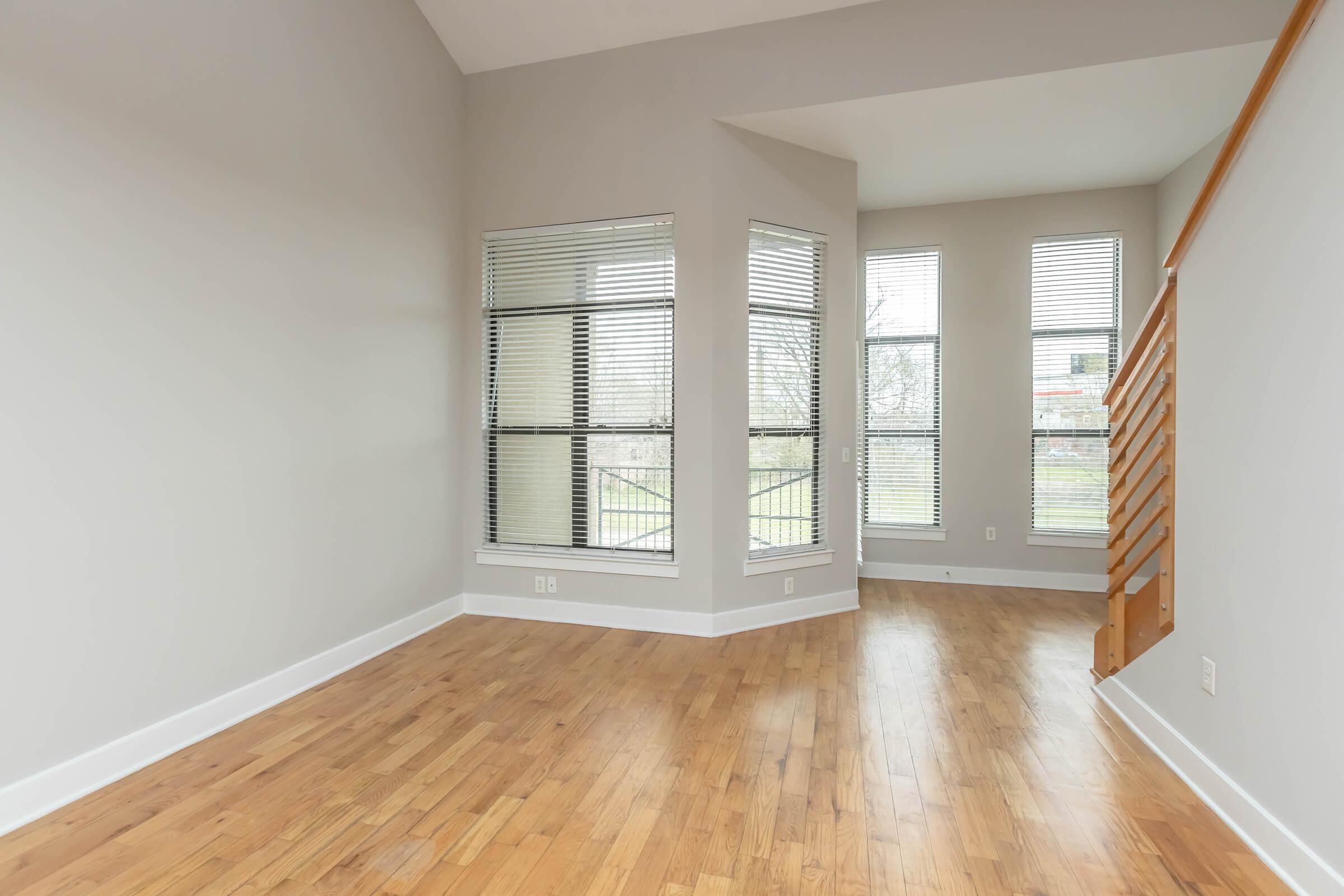 A spacious, well-lit interior room featuring light wood flooring, gray walls, and large windows with blinds. There is a short set of stairs leading to another level, and a corner nook with additional windows, creating a bright, airy atmosphere.