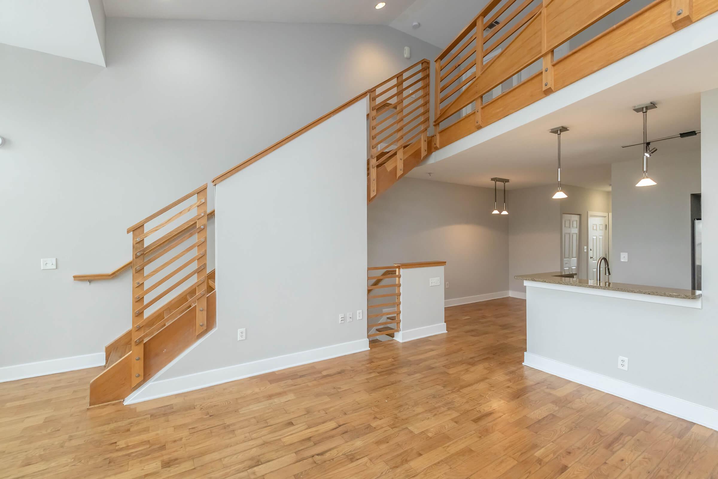 Interior of a modern, open-concept living space featuring a wooden staircase, light gray walls, and hardwood flooring. The area includes a kitchen with a granite countertop and pendant lighting, along with a spacious living area.
