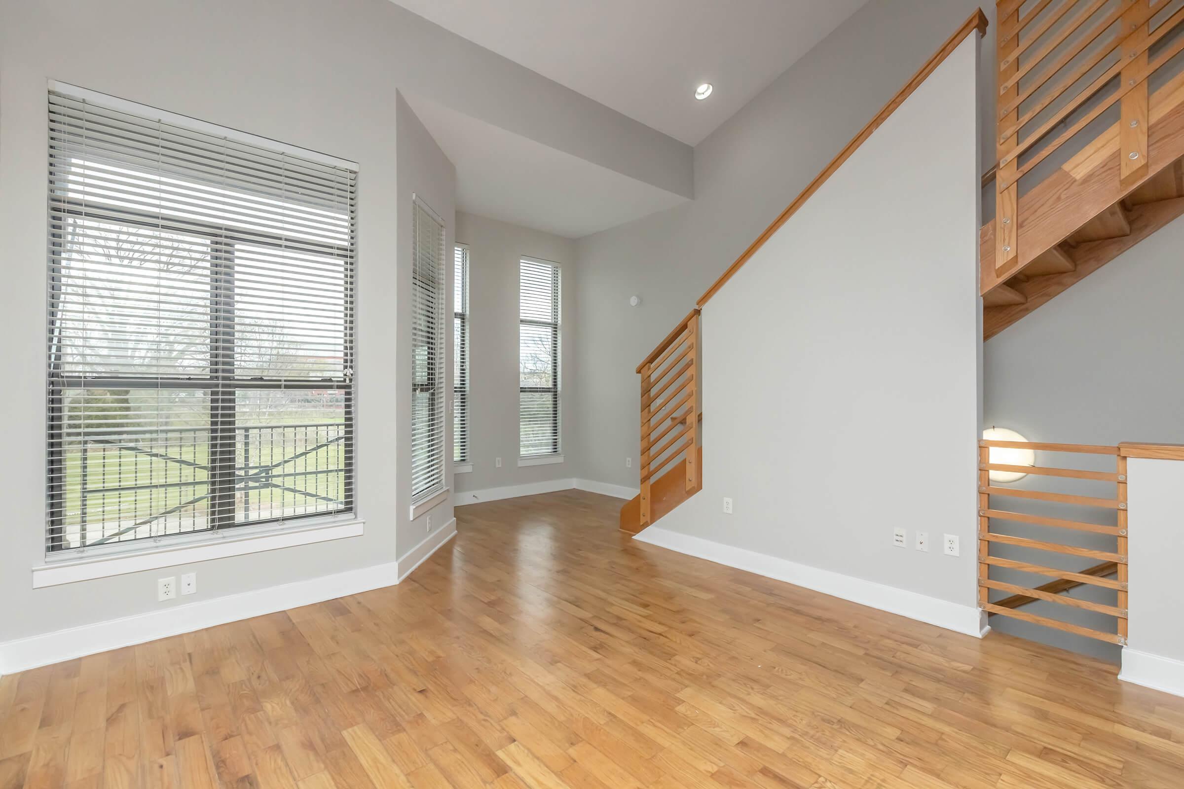 A spacious and modern interior featuring wooden flooring, large windows with blinds, and a staircase with wooden railings. The walls are painted in a light gray color, enhancing the brightness of the space. Natural light fills the area, creating an inviting atmosphere.