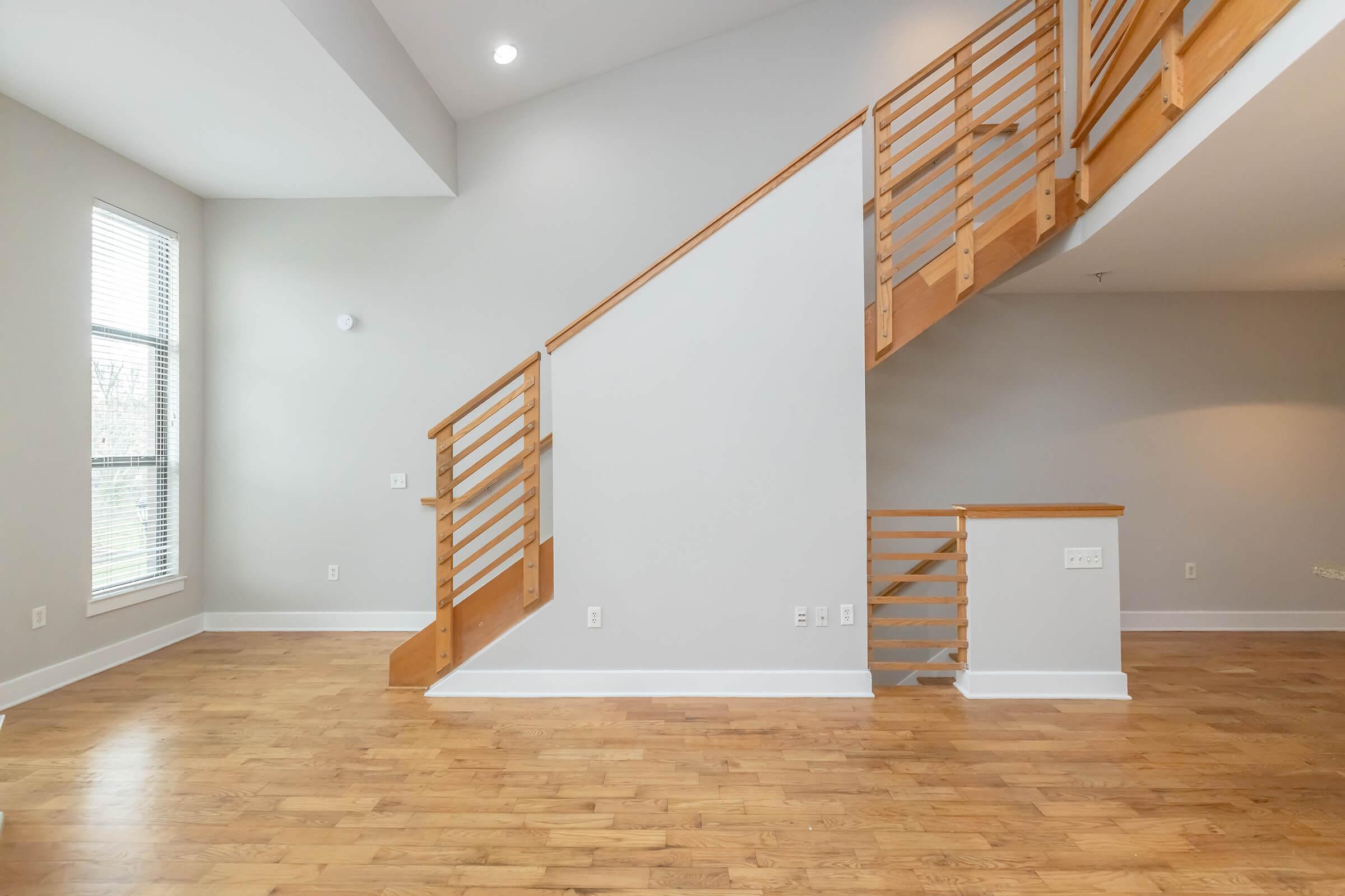 Interior view of a spacious, modern living area featuring light-colored walls and hardwood flooring. A wooden staircase with sleek railings is prominently displayed, leading to a second level. Large windows allow natural light to fill the room, creating a bright and open atmosphere.