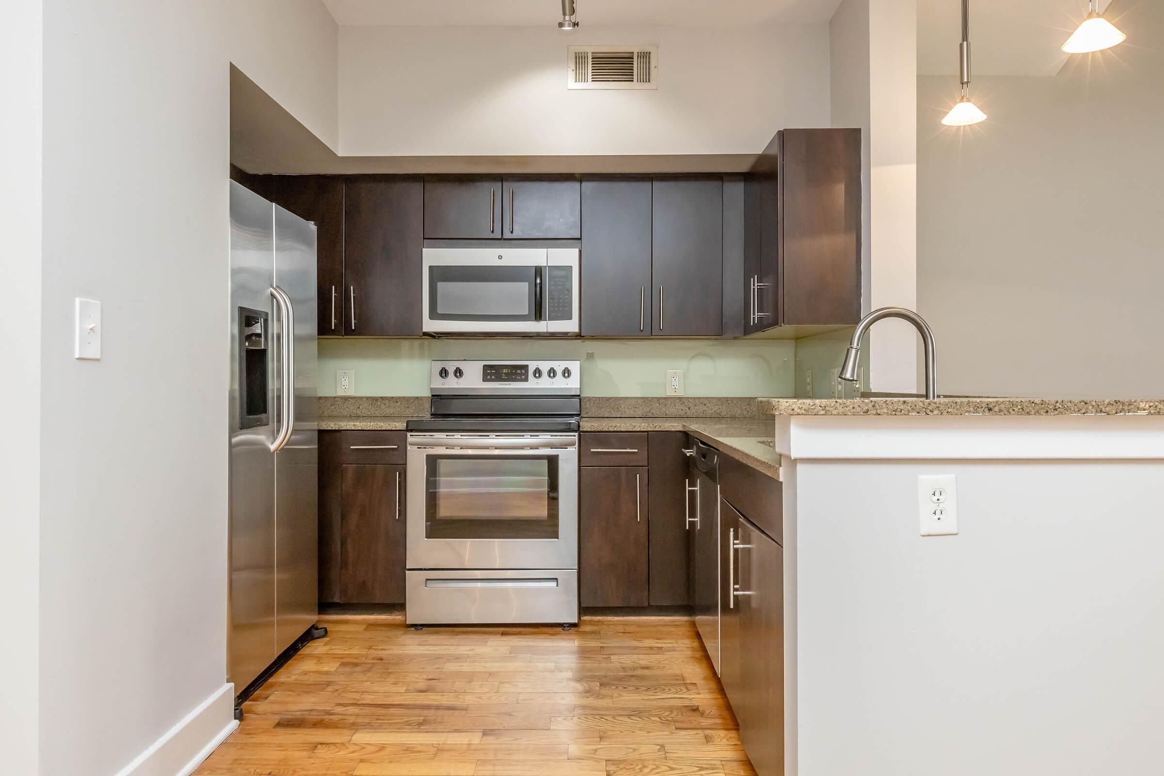 Modern kitchen featuring dark wooden cabinets, stainless steel appliances including a refrigerator, oven, and microwave. The granite countertop complements the sleek design, with pendant lights illuminating the space. Wood flooring adds warmth to the contemporary interior.