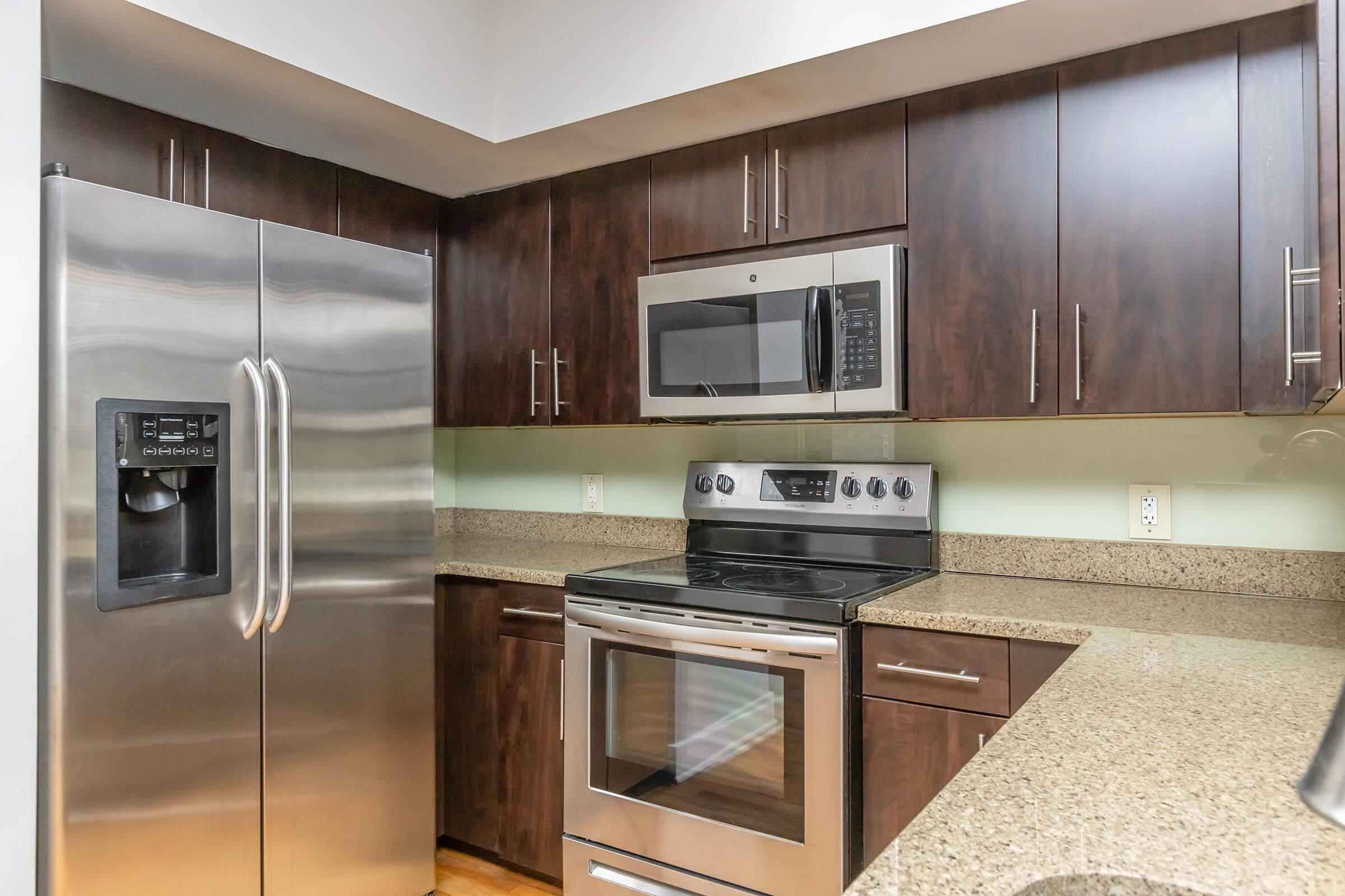 Modern kitchen featuring dark wooden cabinets, stainless steel refrigerator, and microwave. A sleek stove with an oven is integrated into the countertop, complemented by a light-colored granite surface. The overall design is clean and contemporary, enhancing the kitchen's functionality and aesthetic appeal.