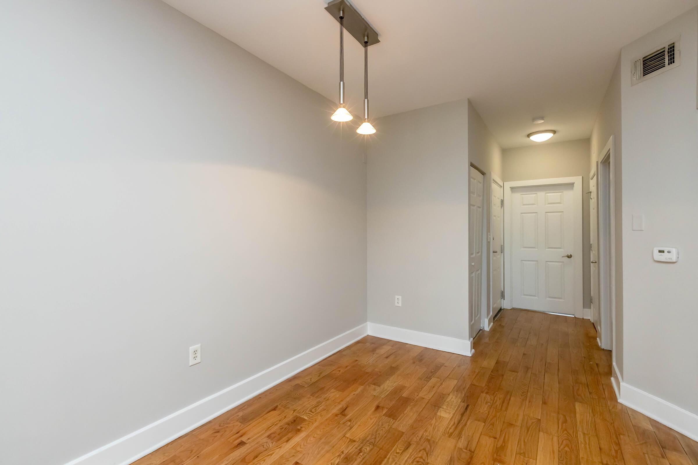 A well-lit interior space featuring light gray walls and polished wooden flooring. Two pendant lights hang from the ceiling, illuminating the area. A hallway with closed doors is visible on the right, leading to other rooms in the building. The overall ambience is clean and modern.