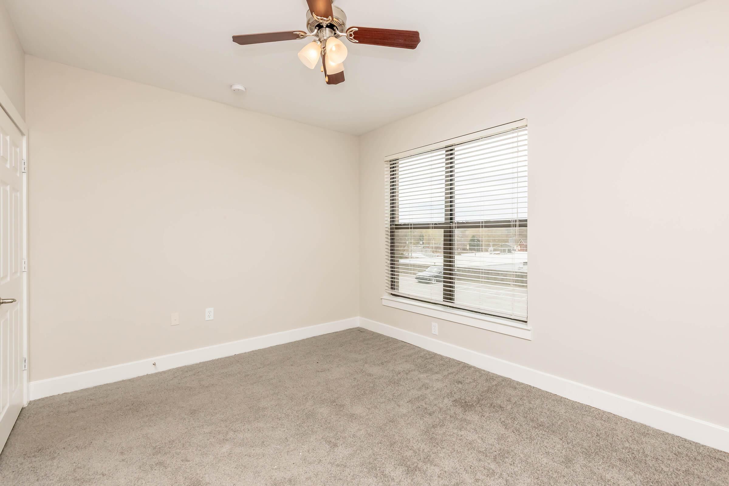A bright, empty room with light gray carpet and a ceiling fan. The walls are a neutral color, and there is a large window with blinds that lets in natural light. The room has a minimalist design, making it versatile for different uses, such as a bedroom or office space.
