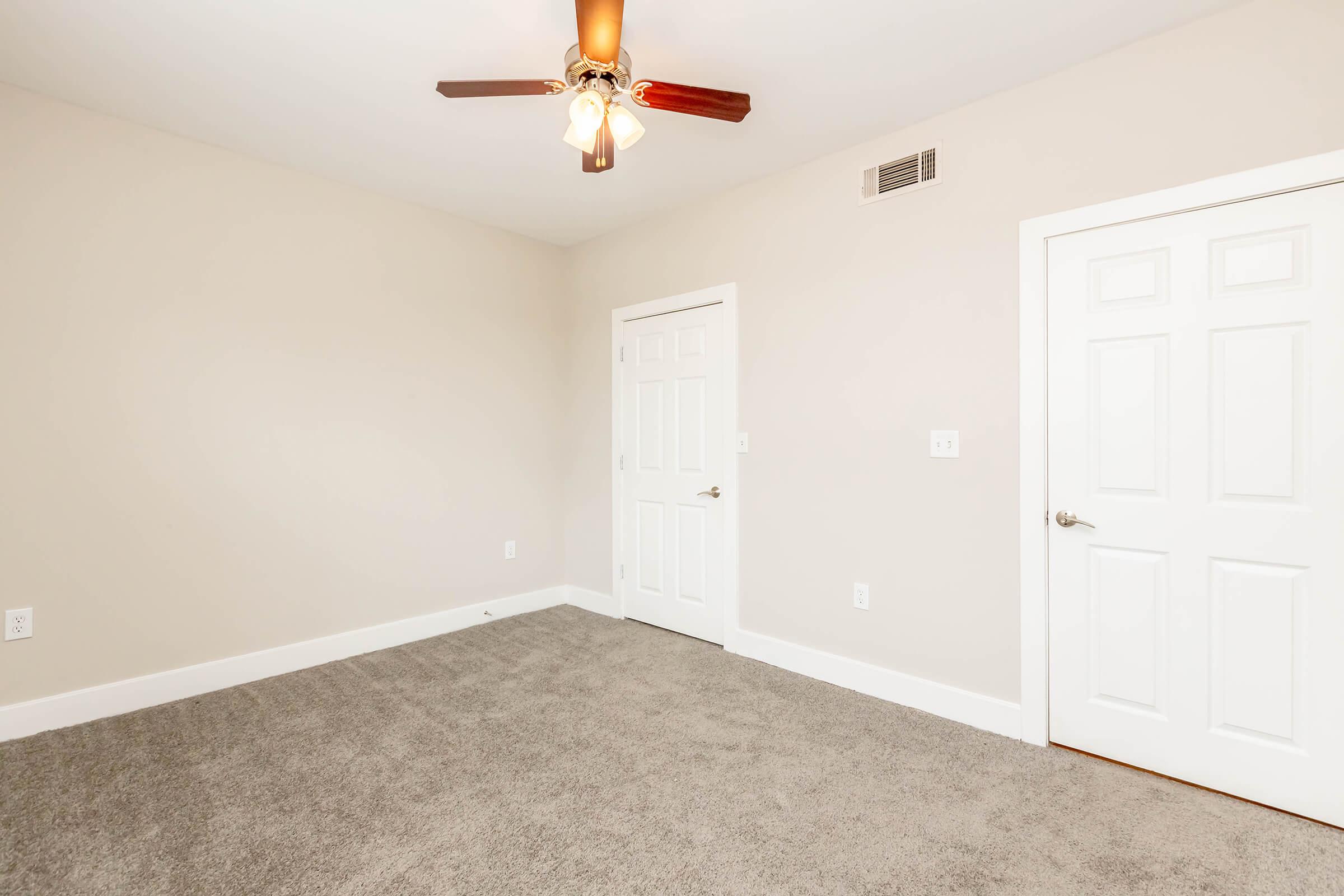 A spacious room with light beige walls and a soft carpet. There is a ceiling fan with wooden blades, and two white doors leading to closets or other areas. Natural light fills the room, creating a calm atmosphere, and the floor is clean and well-maintained.
