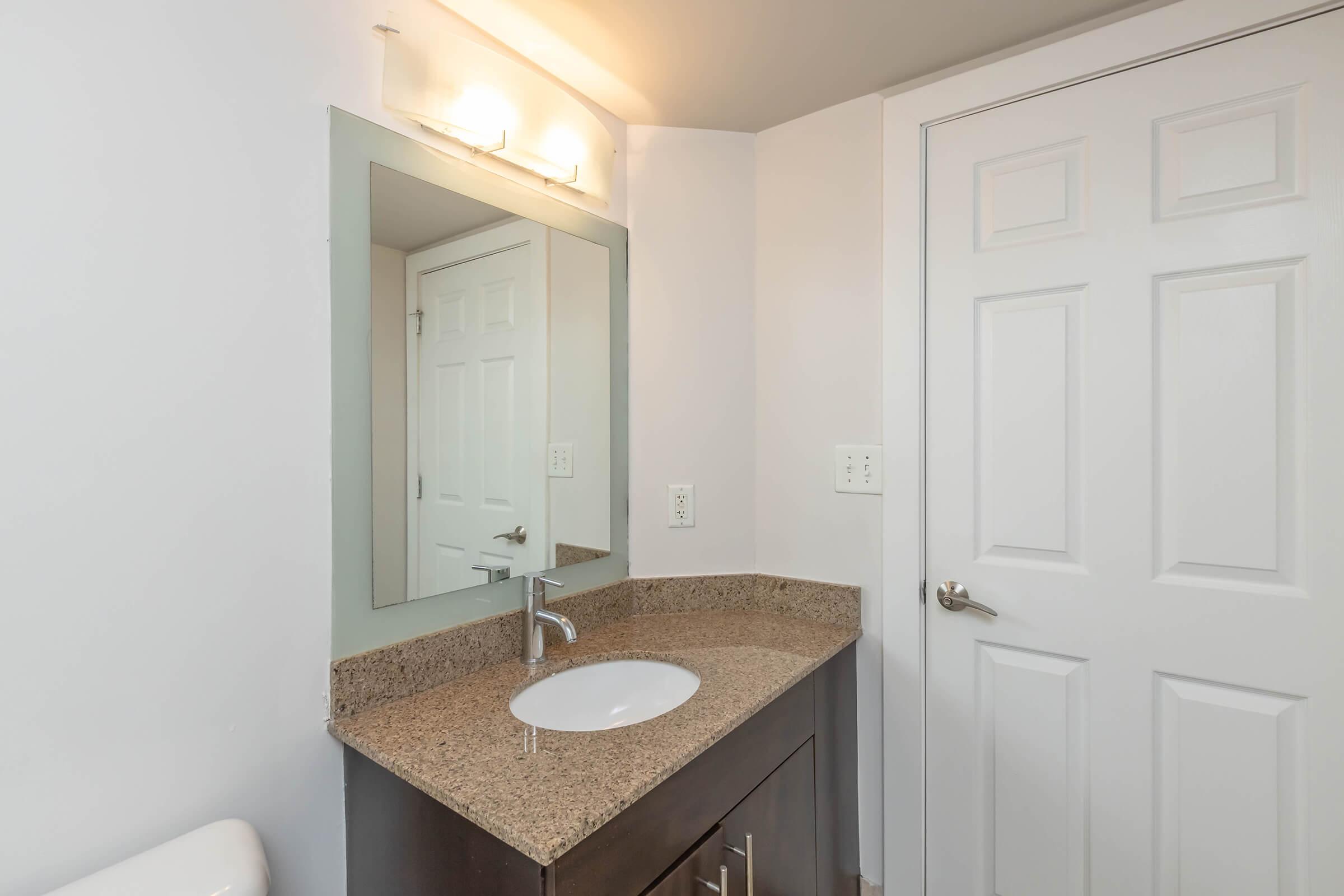A modern bathroom featuring a granite countertop with a sink and a mirror above it. The walls are painted white, and there's a closed white door visible. Soft lighting illuminates the space, creating a bright and clean atmosphere. The flooring is light-colored tile.