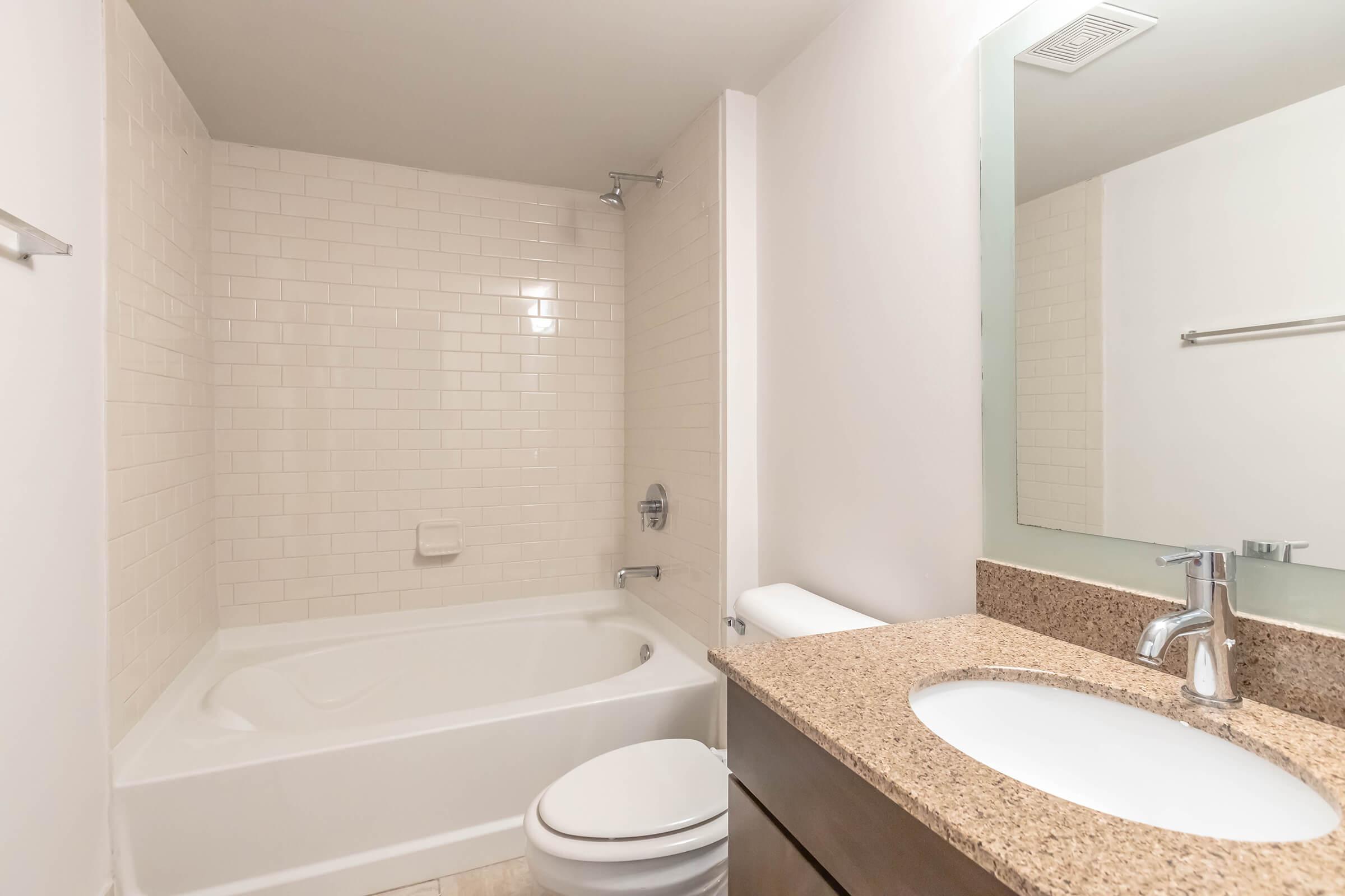 A clean, modern bathroom featuring a bathtub with a shower, a toilet, and a vanity with a round sink. The walls are tiled with white subway tiles, and the countertop is made of speckled granite. A large mirror above the sink reflects the light, creating a bright atmosphere.
