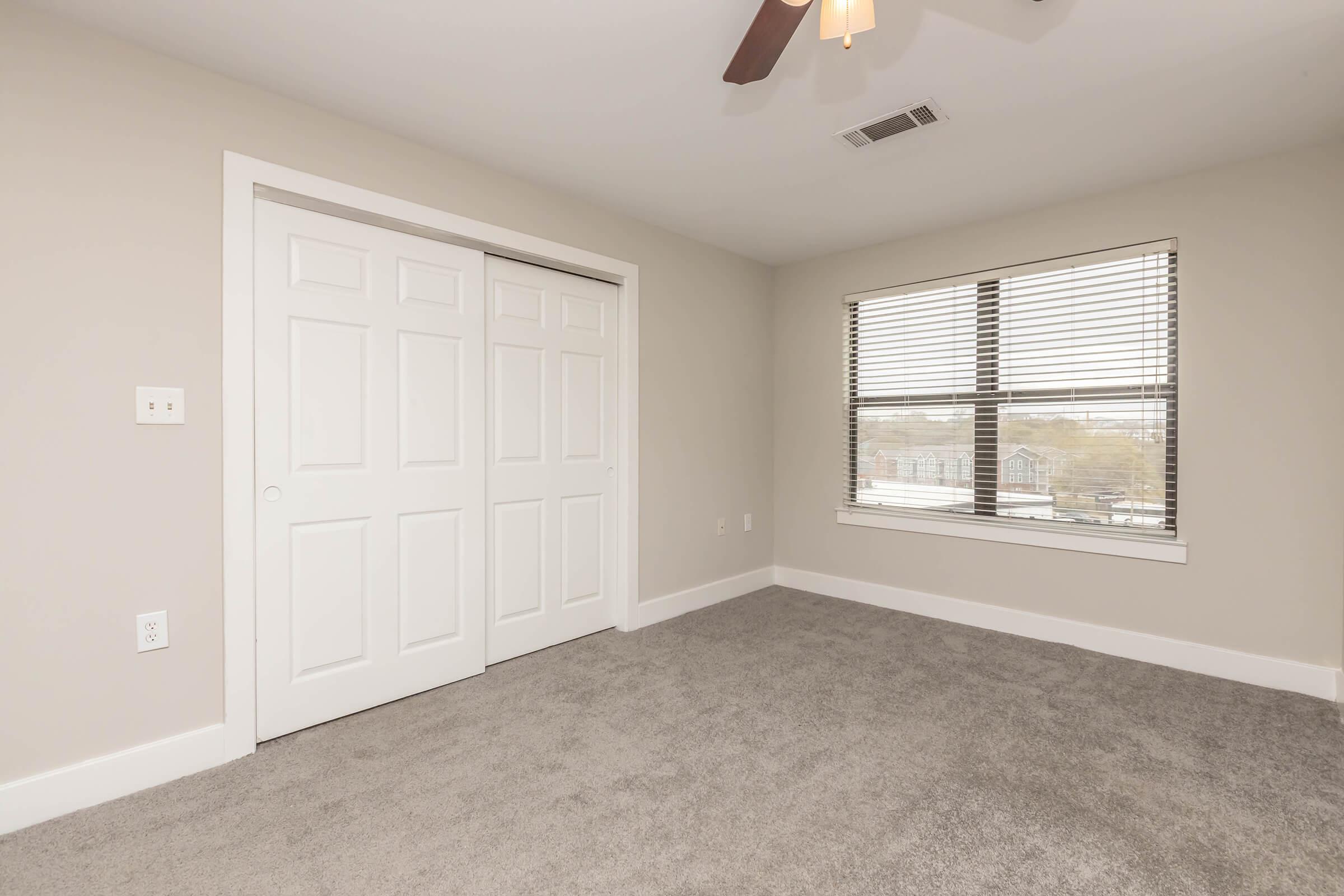 A well-lit bedroom featuring light gray walls, a ceiling fan, and a plush carpeted floor. The room includes a large window with blinds, providing natural light, and a set of white double doors leading to a closet. The overall atmosphere is clean and modern, suitable for personal use.