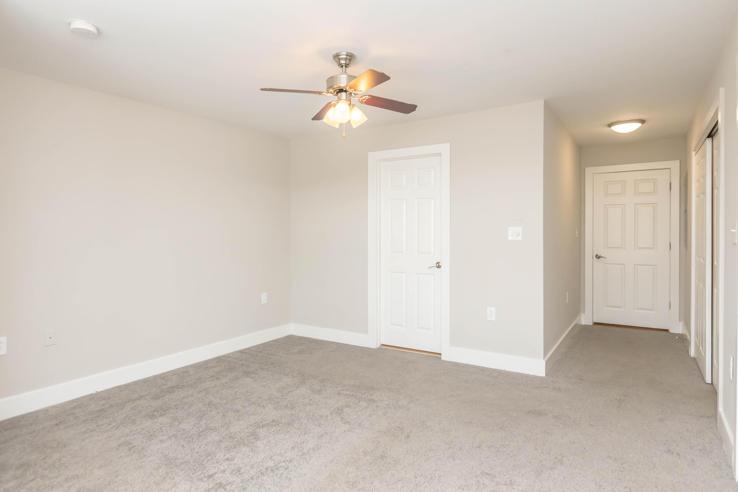 Empty room with light gray walls and a ceiling fan. The floor is covered in light beige carpet. There is a closed white door on the left and a hallway visible at the far end. The room is well-lit, creating a spacious atmosphere, with baseboards around the walls.