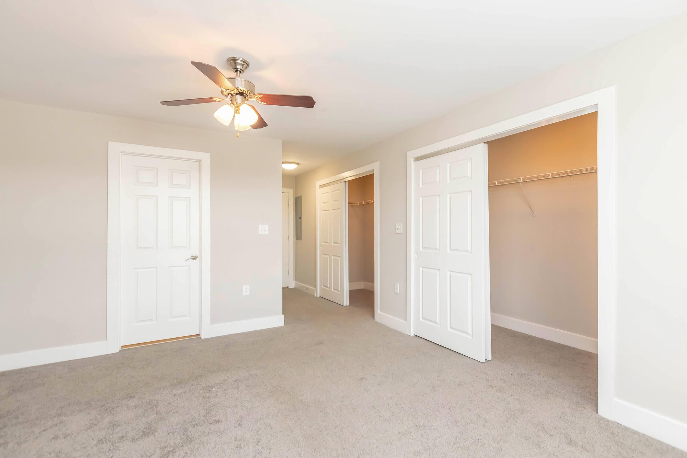 A sparsely furnished room featuring a light-colored carpet and a ceiling fan. The space includes two open doorways leading to other areas and a closet with shelving. The walls are painted a soft beige, creating a bright and airy atmosphere.