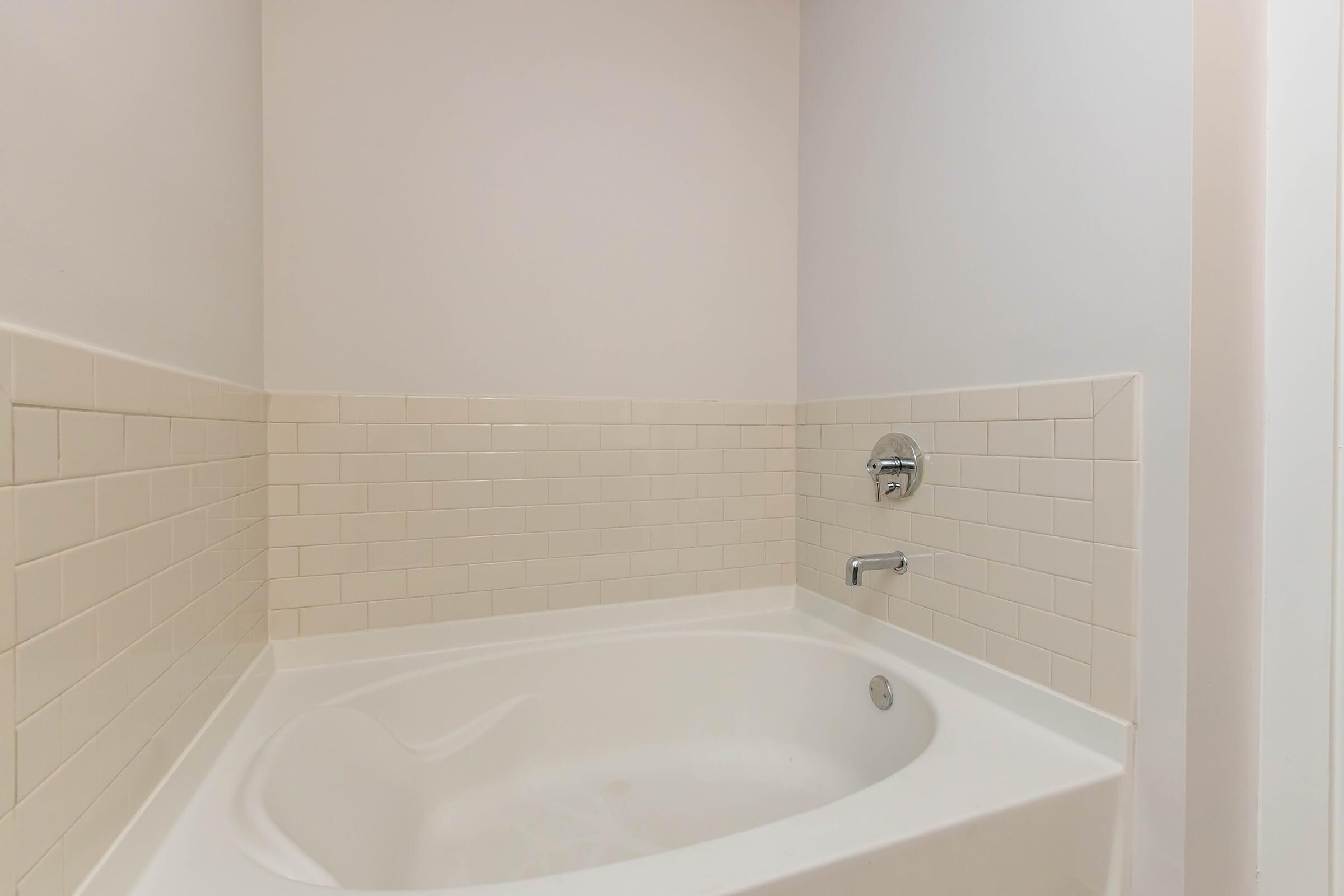 A clean, modern bathroom featuring a spacious white bathtub with a curved design. The surrounding walls are tiled in light-colored square tiles, and there is a chrome faucet installed. The setting has minimal decor, contributing to a calm and inviting atmosphere.