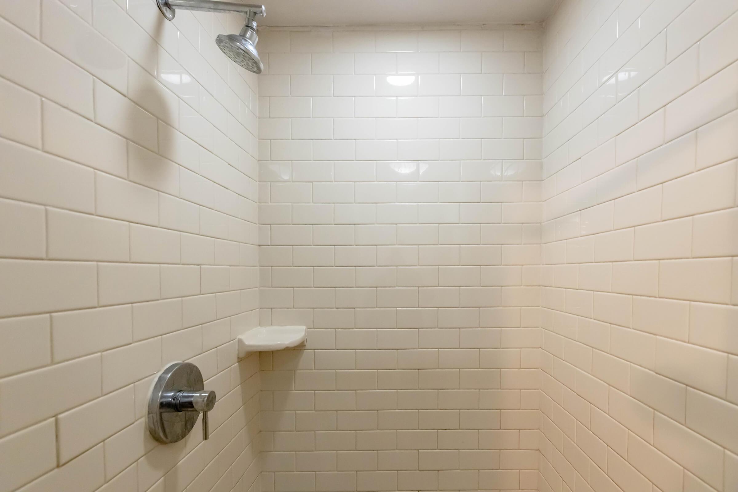 A clean, empty shower space with white tiled walls. There are two showerheads mounted on the wall, a small shelf for toiletries, and bright overhead lighting illuminating the area.