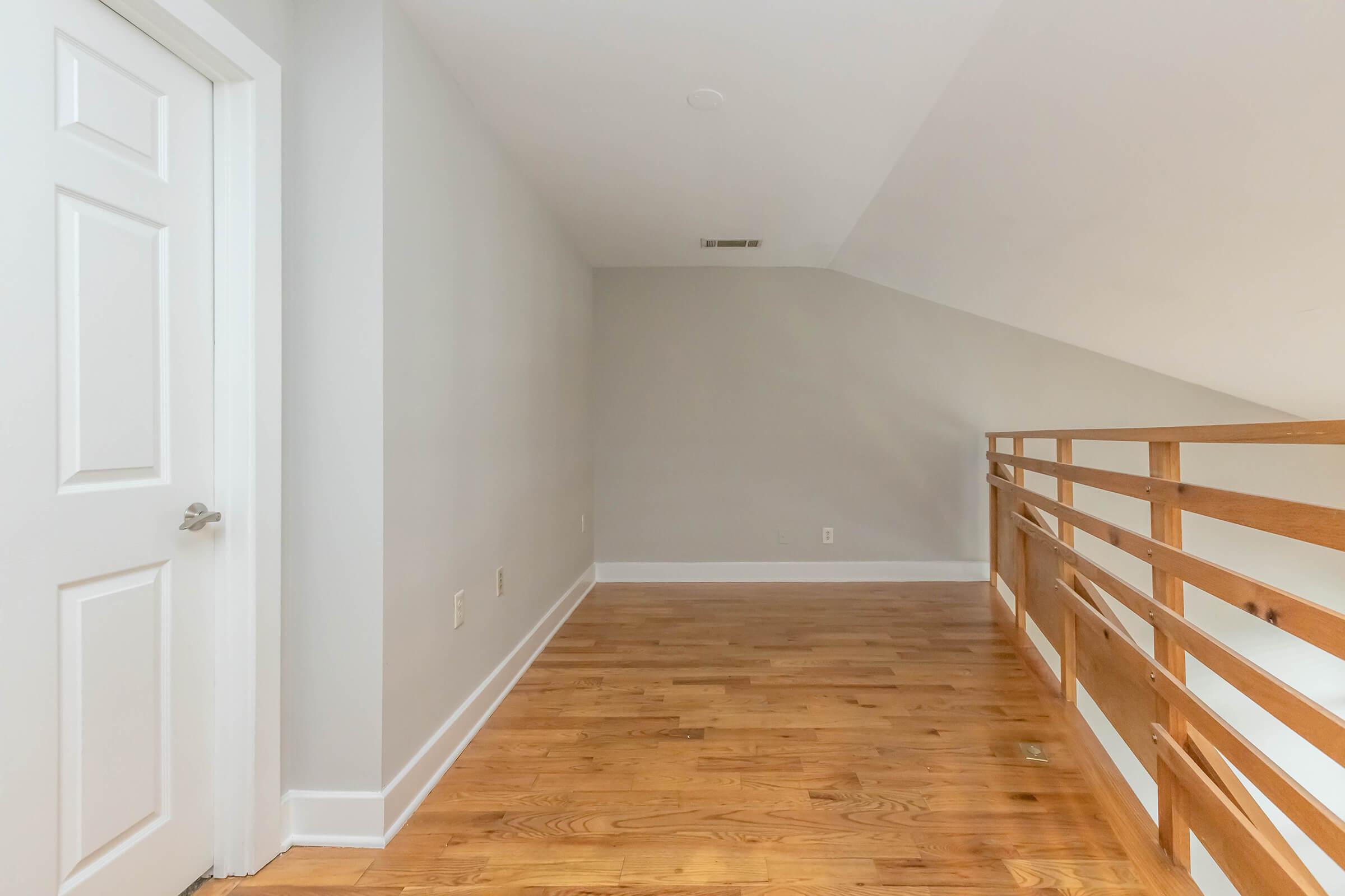 A spacious, empty loft area featuring a wooden railing and hardwood flooring. A white door is visible on the left, and the wall is painted light gray. The ceiling is sloped, creating a cozy atmosphere. Natural light brightens the space, enhancing its openness.