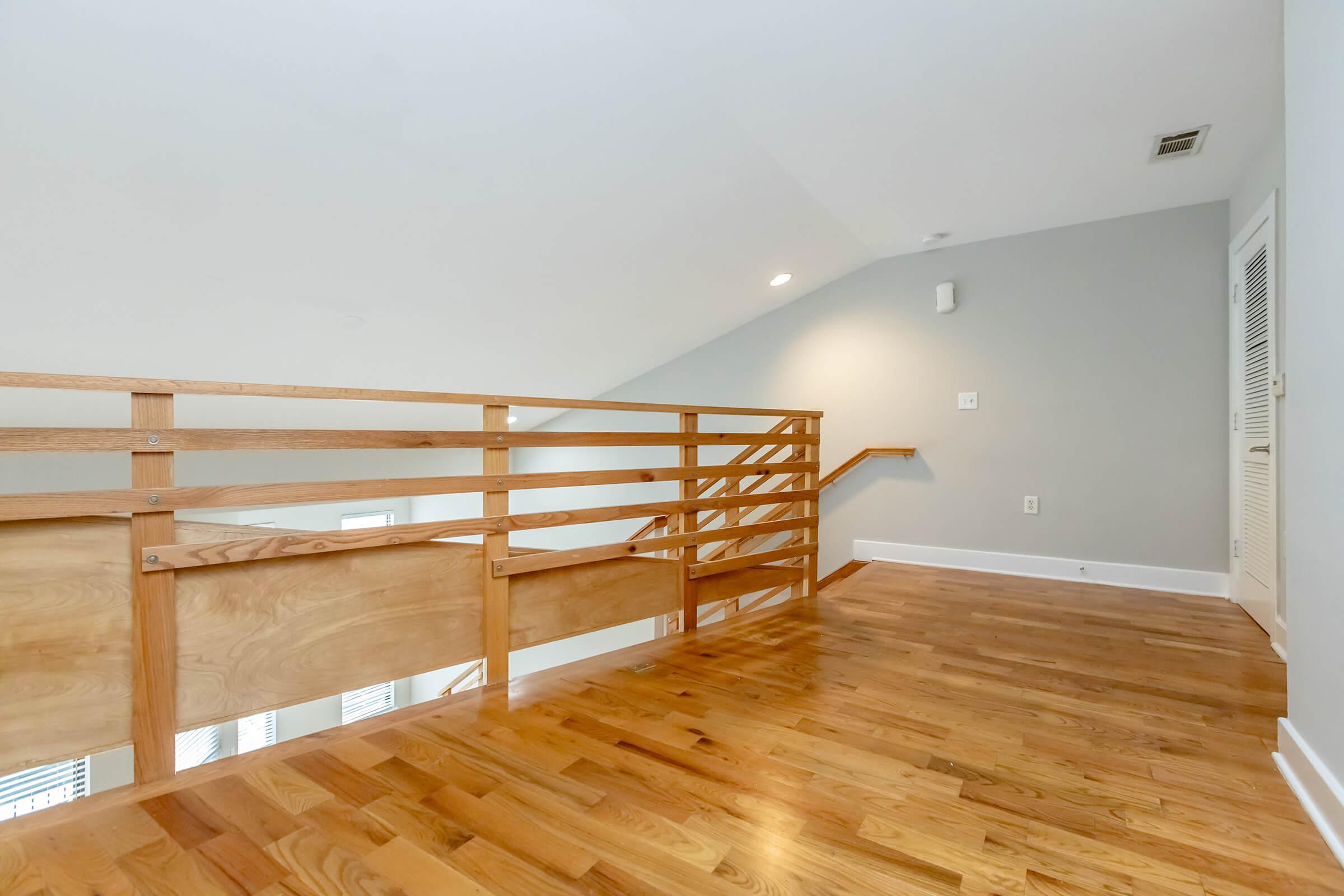 A spacious, well-lit loft area featuring hardwood flooring, a wooden railing, and light gray walls. The design is modern and minimalistic, with natural light flooding in from windows on one side. The space is free from clutter, giving it a clean and open atmosphere.