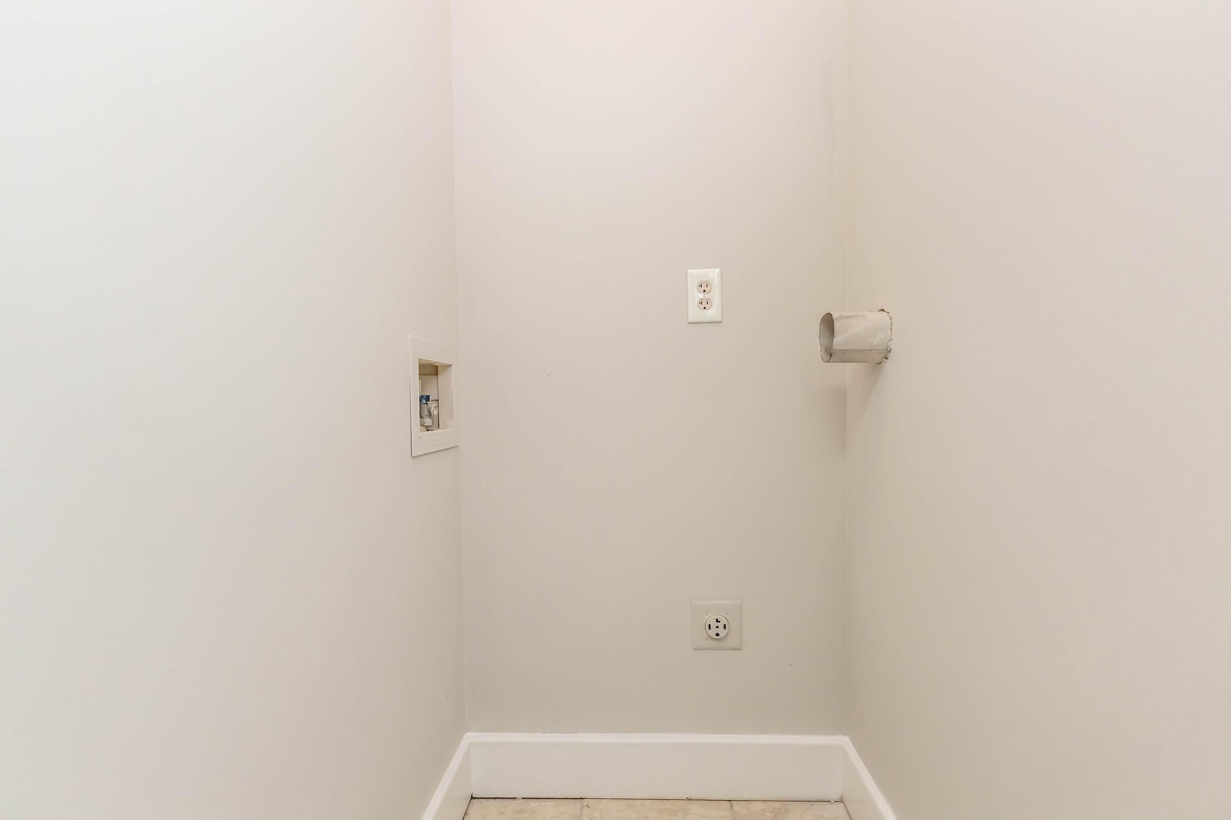 A blank corner of a room with plain white walls. On the left side, there is a small cutout in the wall, and on the right side, two electrical outlets are visible. A vent pipe exits the wall to the right of the outlets, with tiled flooring below. The space appears empty and unfinished.